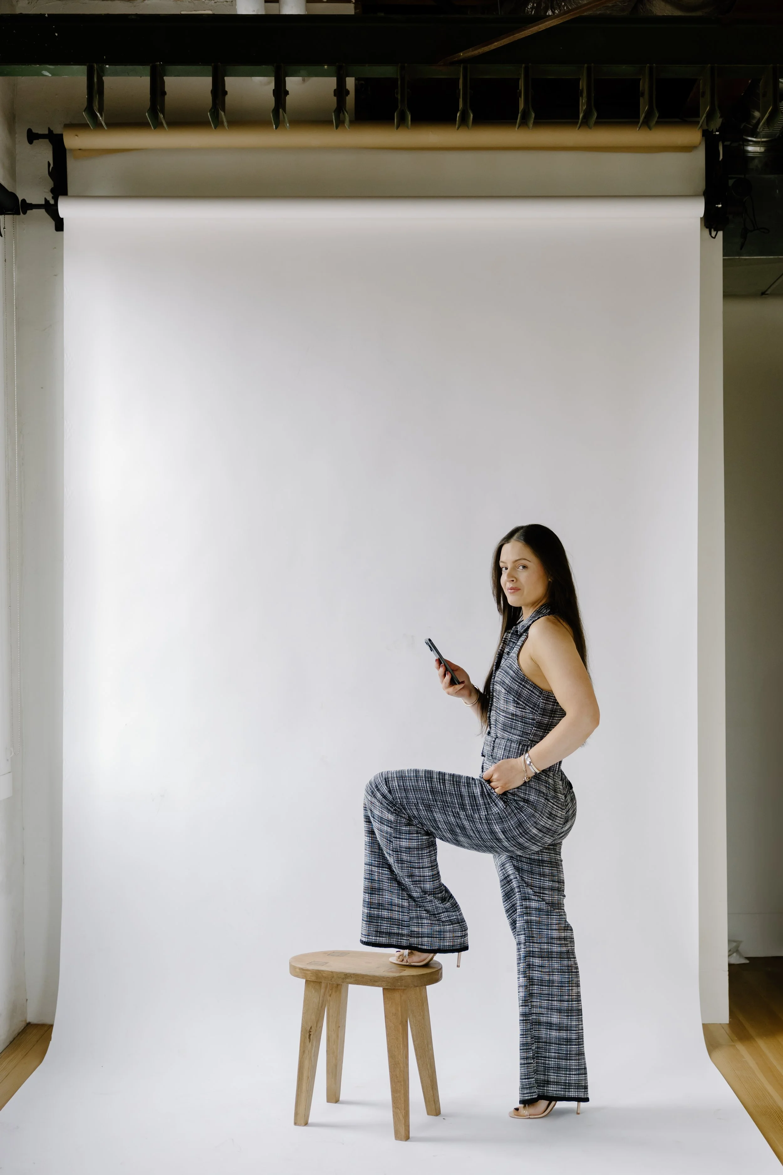 A woman standing on one leg on a wooden stool, with her other foot on her hip, holding a smartphone in her right hand, in front of a white photography backdrop in a studio setting.