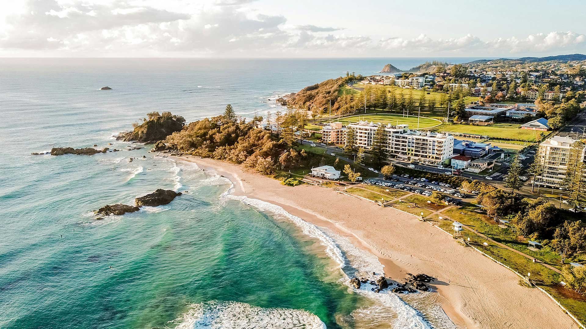 Aerial view of a beach with turquoise water, sandy shoreline, rocks, and greenery. Behind the beach are residential buildings, a park, and hilly terrain with trees.