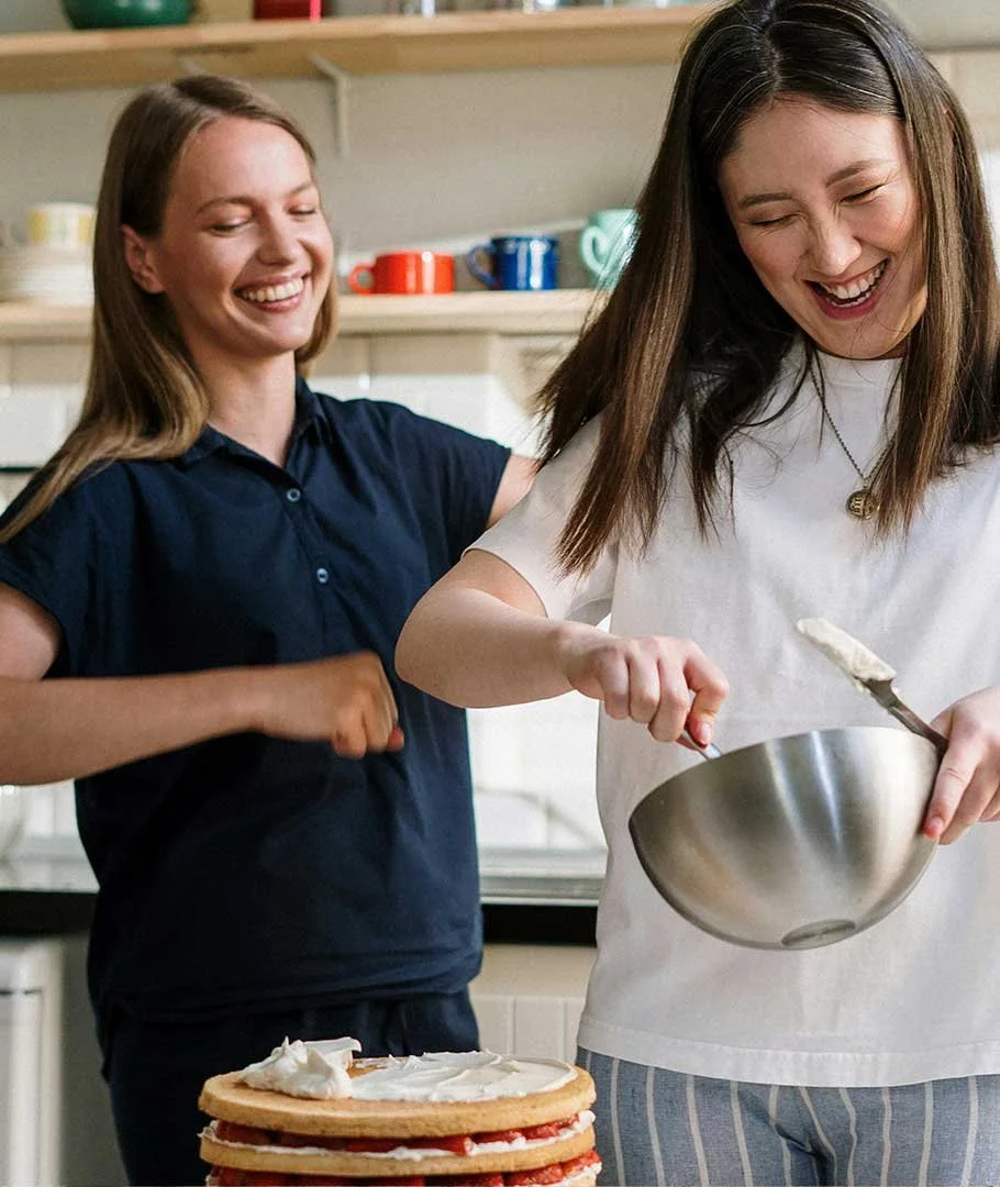 Two young women smiling and cooking together in a kitchen, with one spreading whipped cream or frosting on a layered cake and the other standing nearby.
