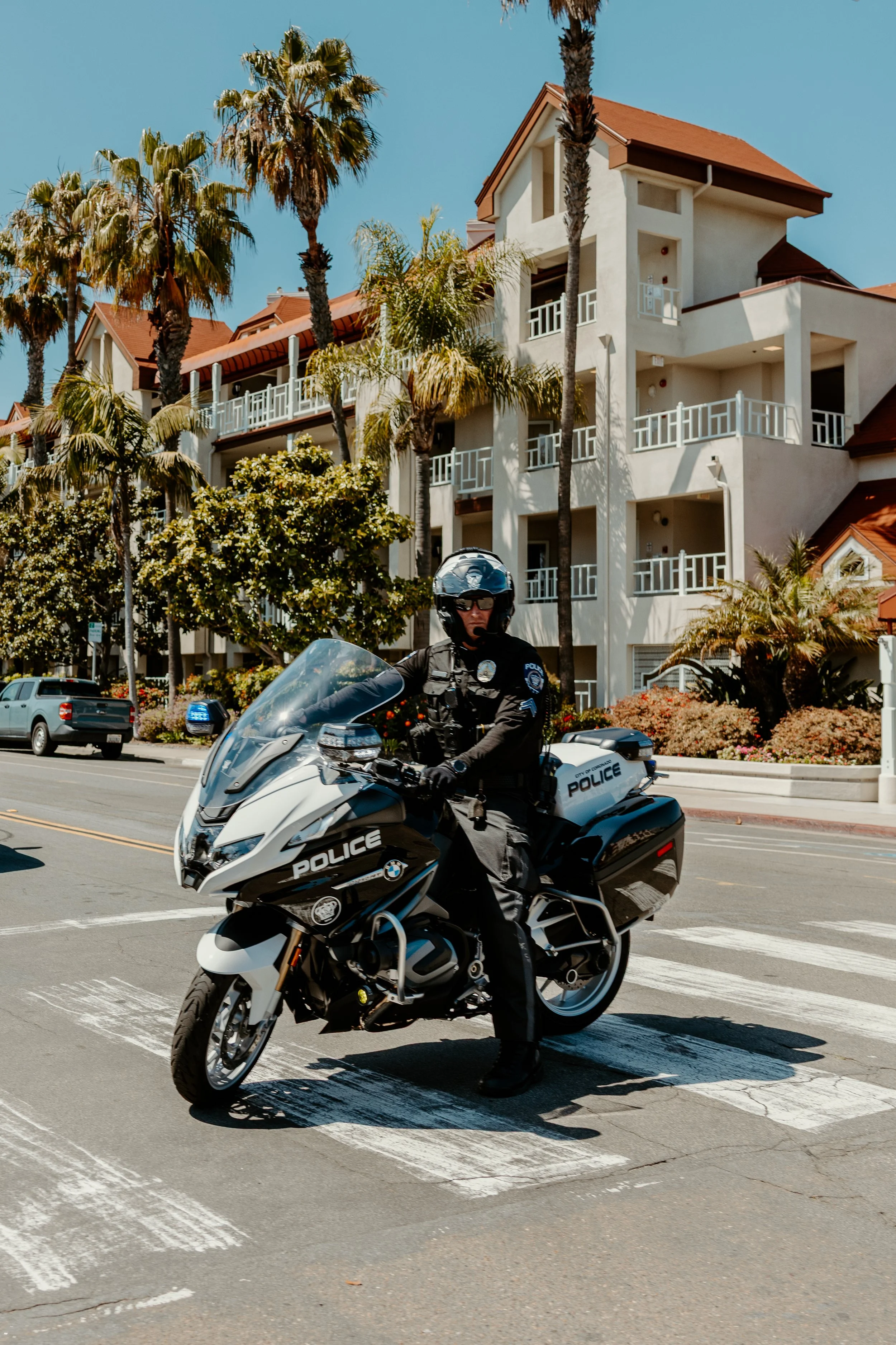 A police officer on a motorcycle stopped at a crosswalk on a city street with hotel buildings and palm trees in the background.