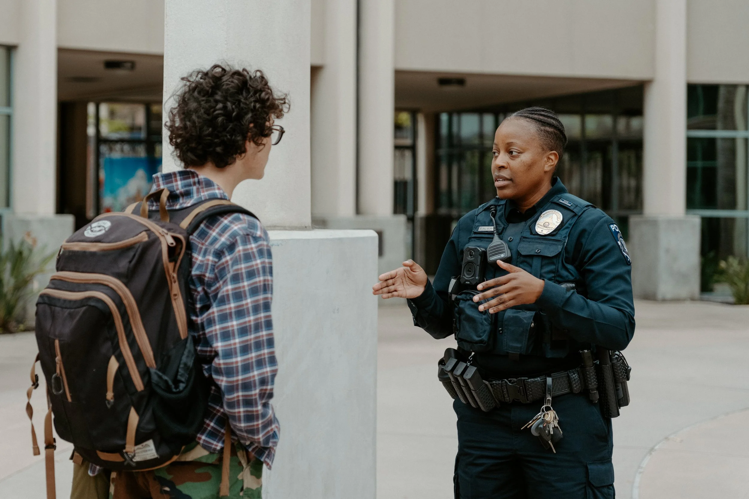 A young person with curly hair wearing glasses, a plaid shirt, and camouflage pants, talking to a female police officer outside a modern building.