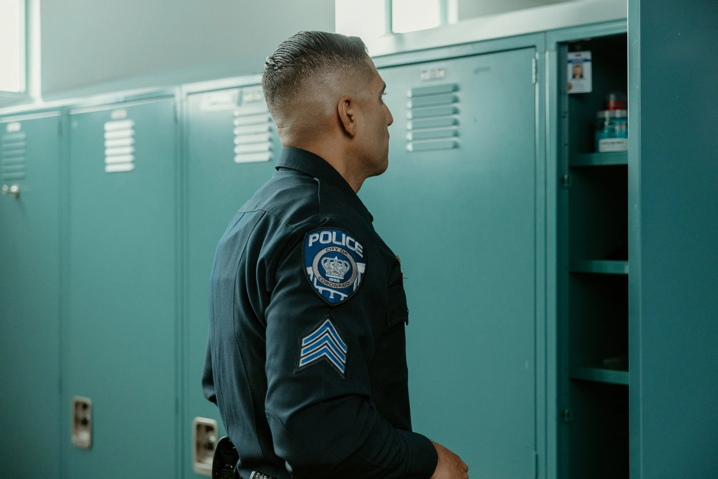 A police officer in uniform standing in front of a row of teal lockers, with one locker door open and items inside.