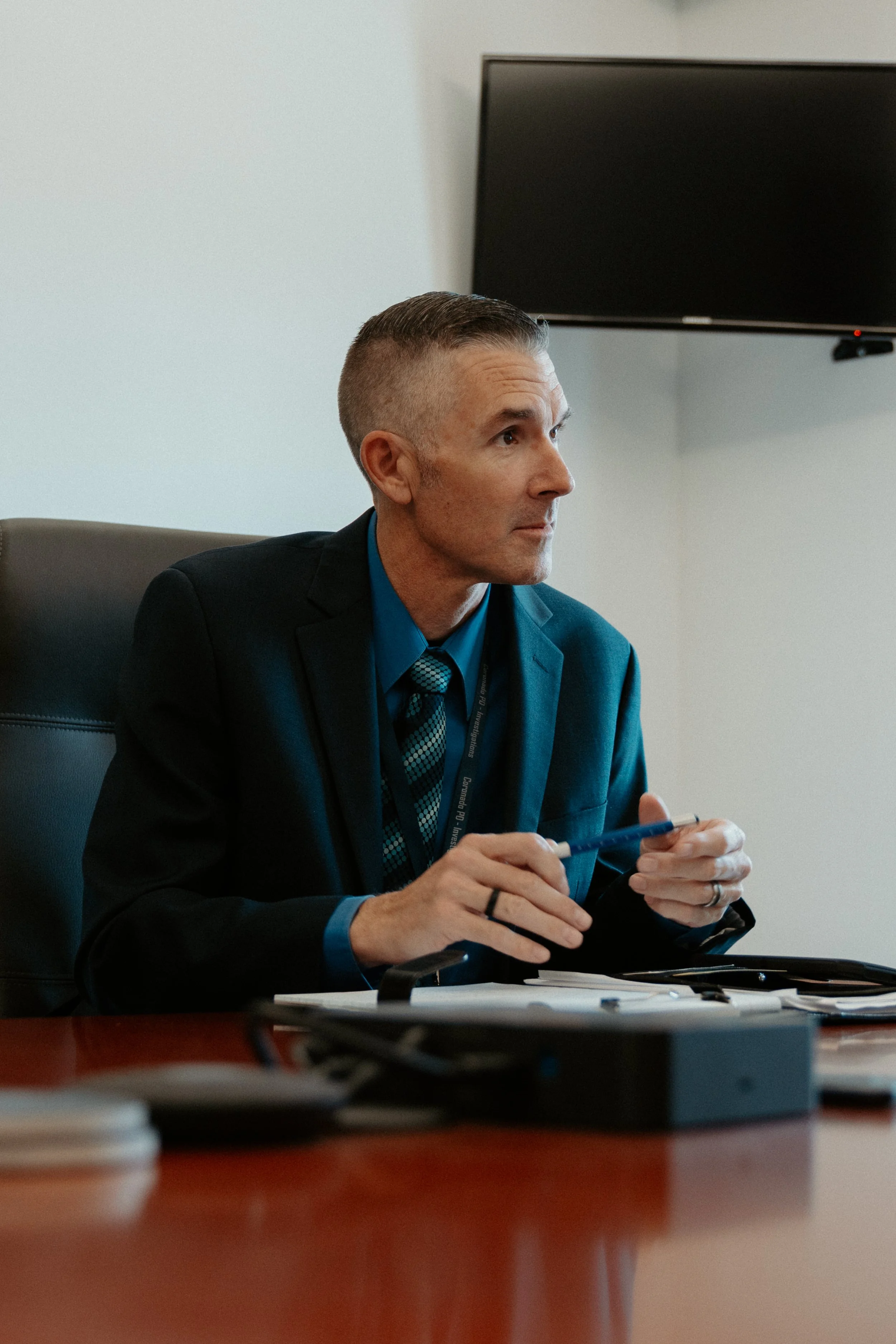 A man in a business suit sitting at a desk in a conference room, holding a pen and looking attentively to the side. There is a large monitor on the wall behind him.