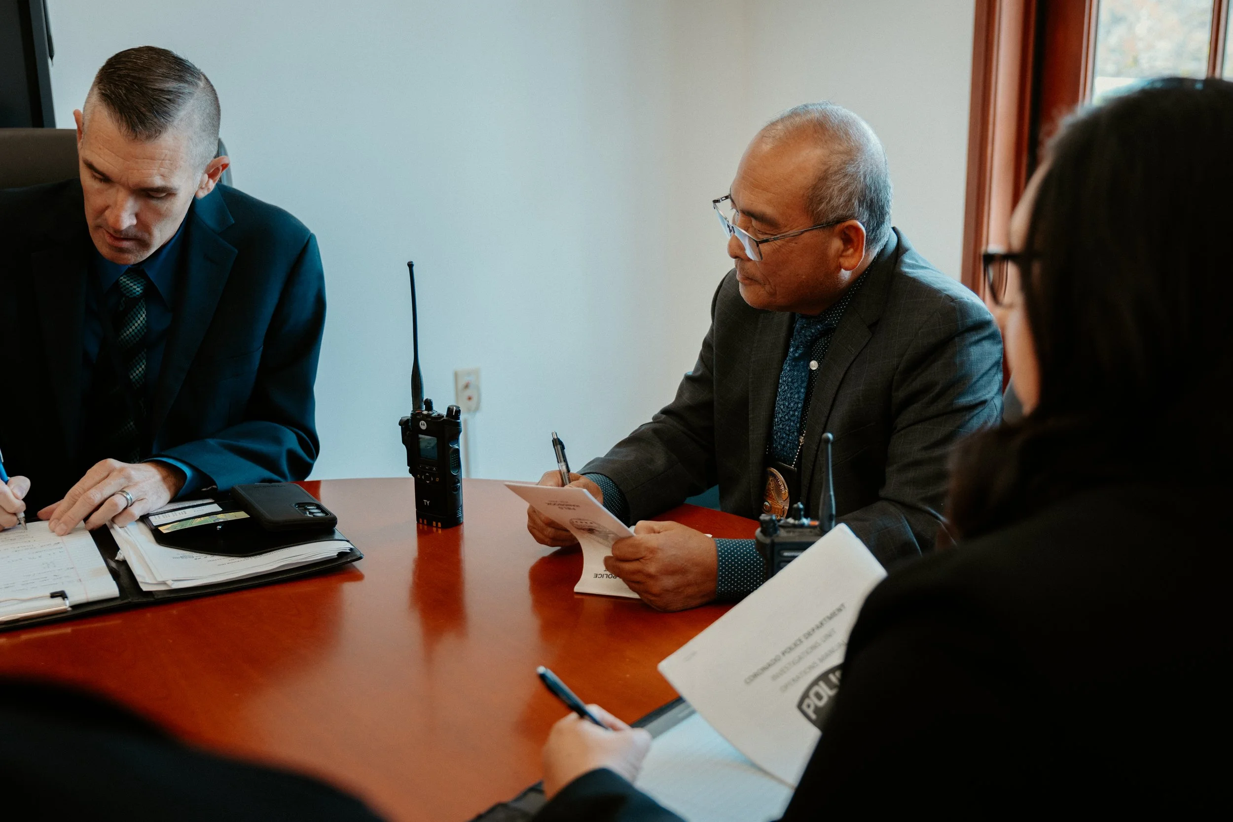 Four people in a meeting room, three dressed in professional suits, with notebooks, pens, and communication devices on the table.