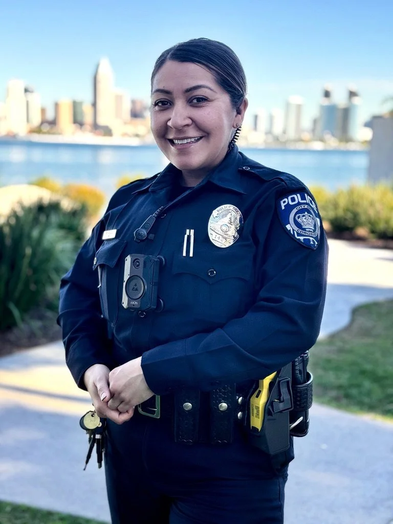 A smiling female police officer in uniform standing outdoors with a city skyline and river in the background.