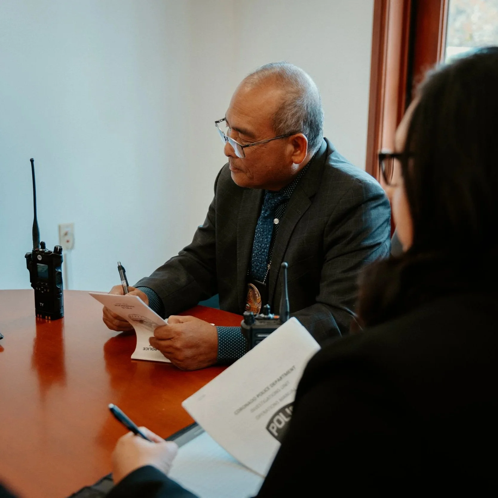 A man in a business suit and glasses sitting at a table, reading a document, with two police radios on the table, and a woman next to him taking notes.