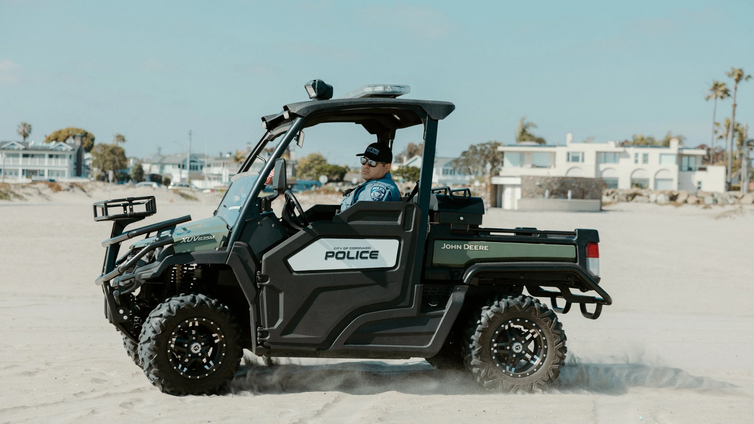A police officer driving a black and green John Deere UTV on a sandy beach with houses and palm trees in the background.