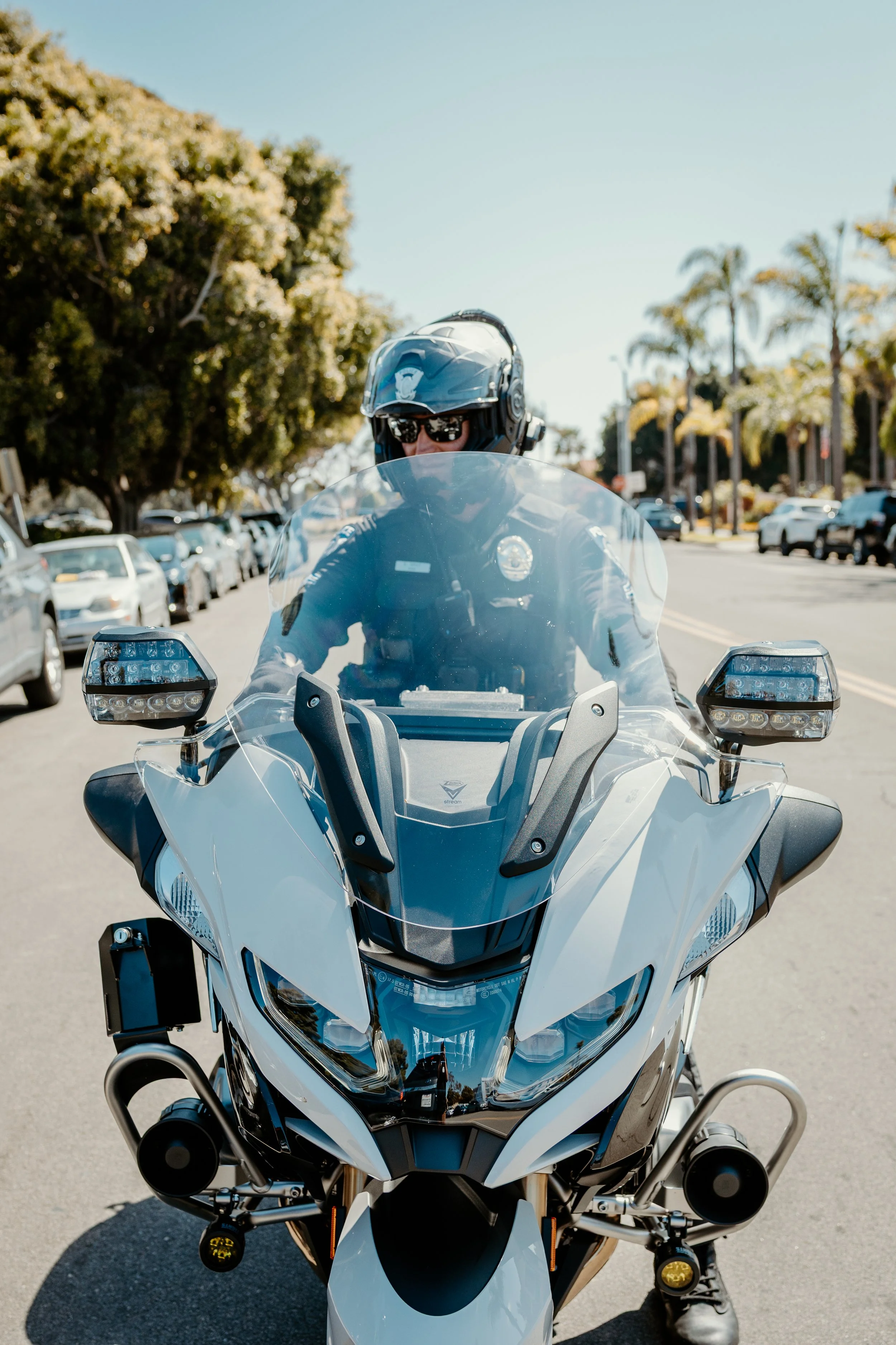 Front view of a person riding a white police motorcycle, wearing a helmet and sunglasses, on a city street with parked cars and trees in the background.