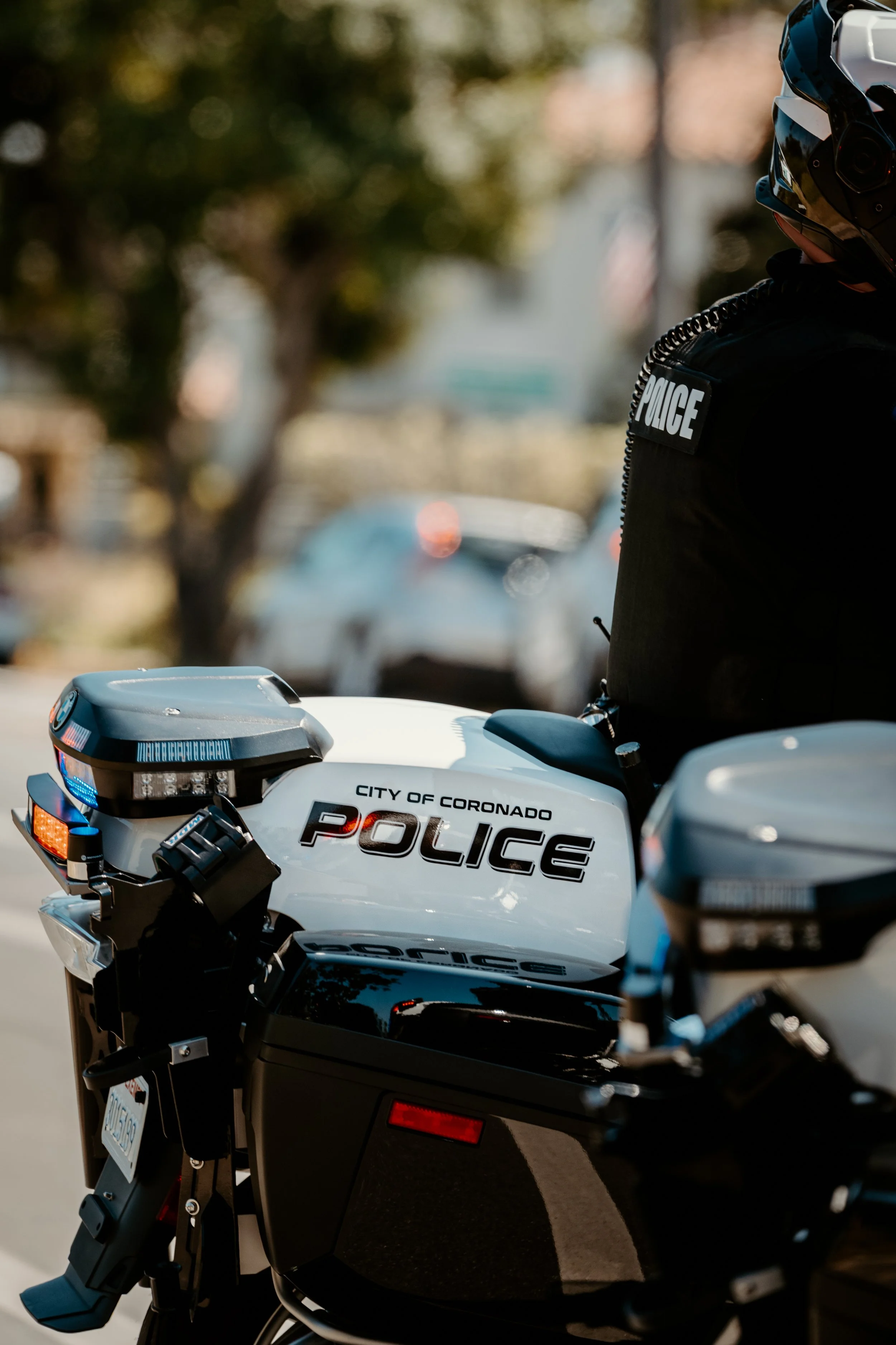 Close-up of a police motorcycle with police officer standing beside it, in a city street with trees and cars in the background, during daytime.
