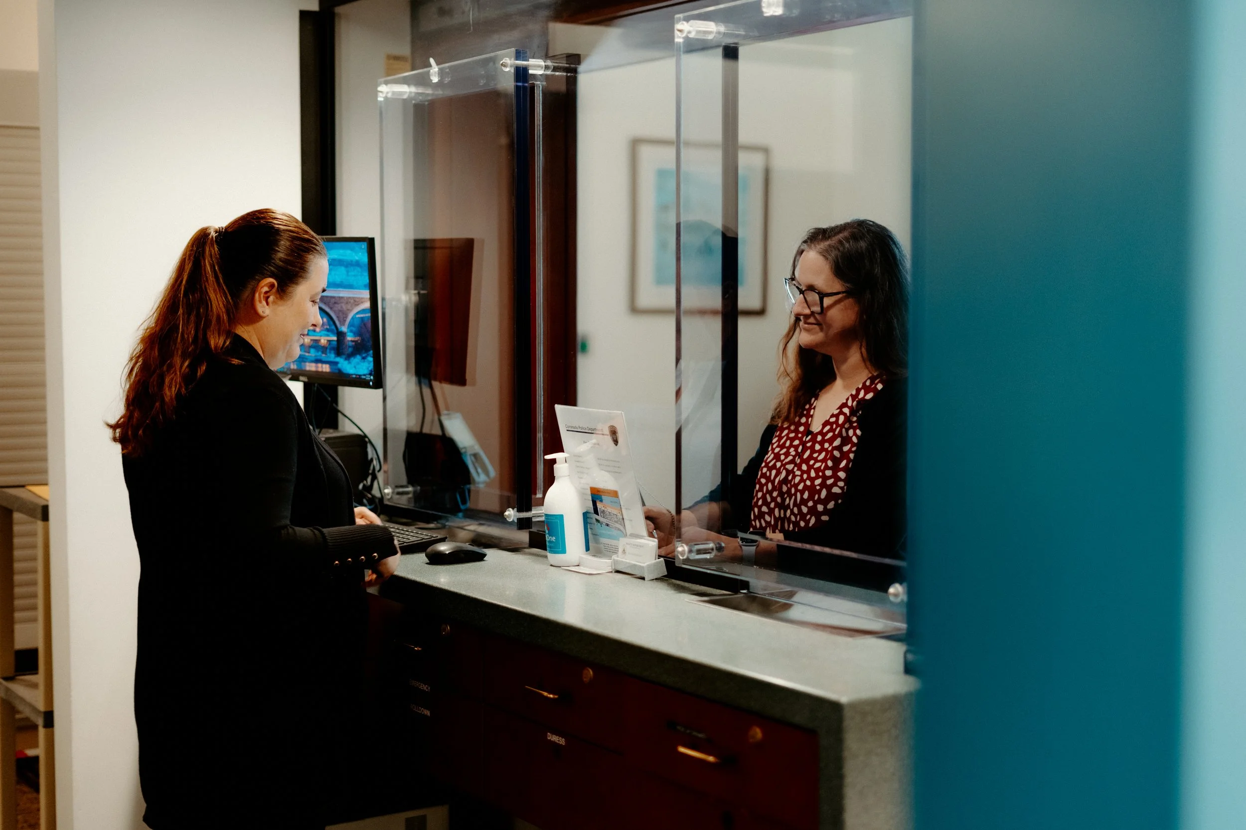 A woman at a reception desk talking to another woman behind a plastic divider in an office setting.