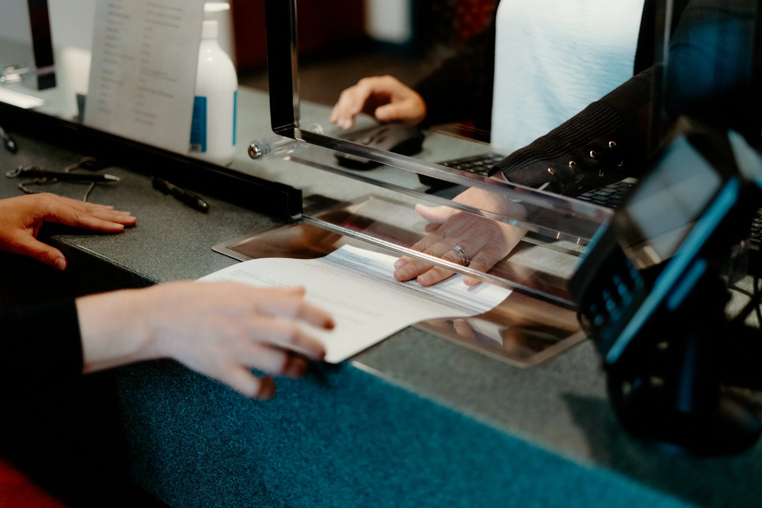 A person using a glass counter to check an ID while an employee handles a document at a reception desk.