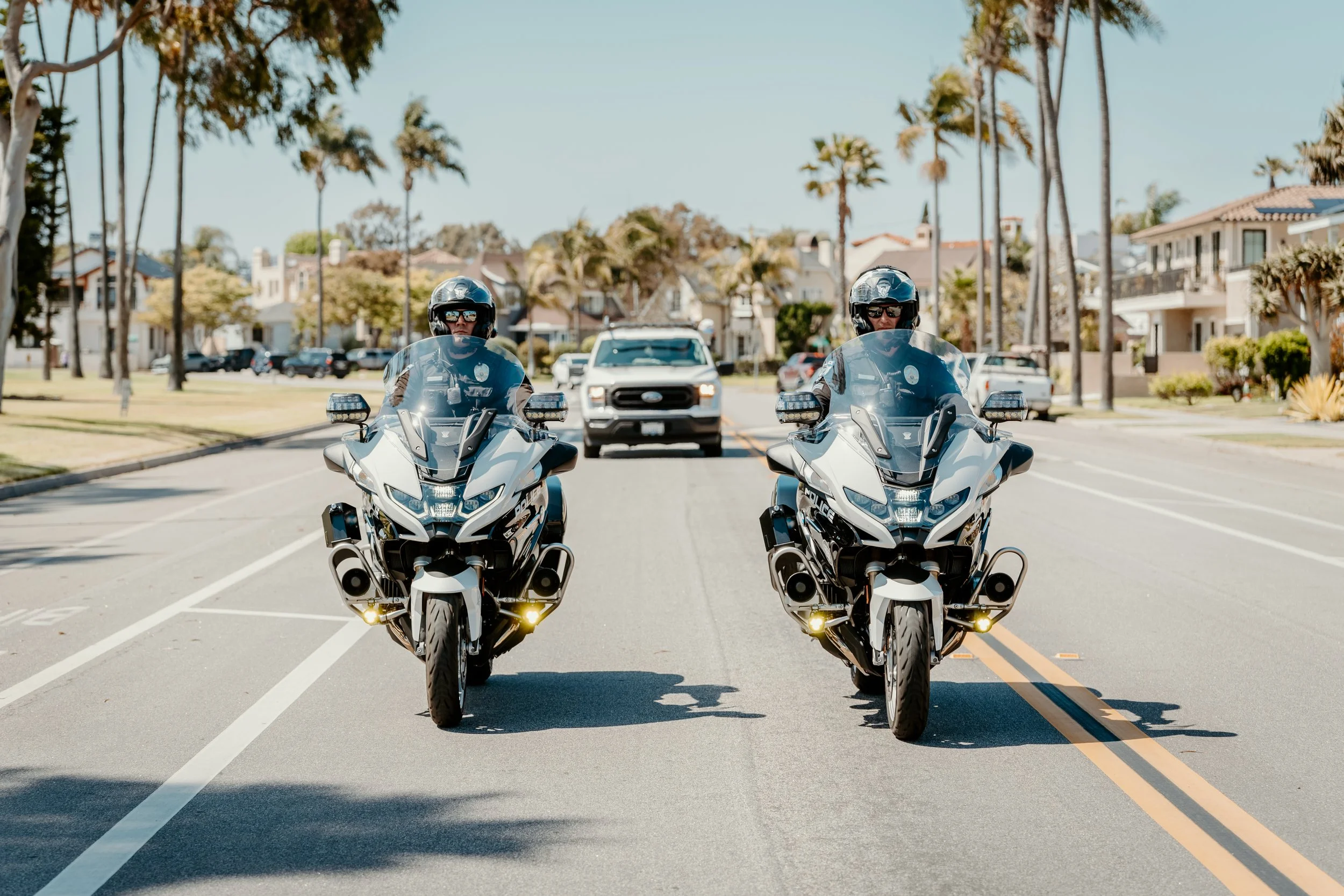 Two police officers riding motorcycles on a city street with palm trees and residential houses in the background.