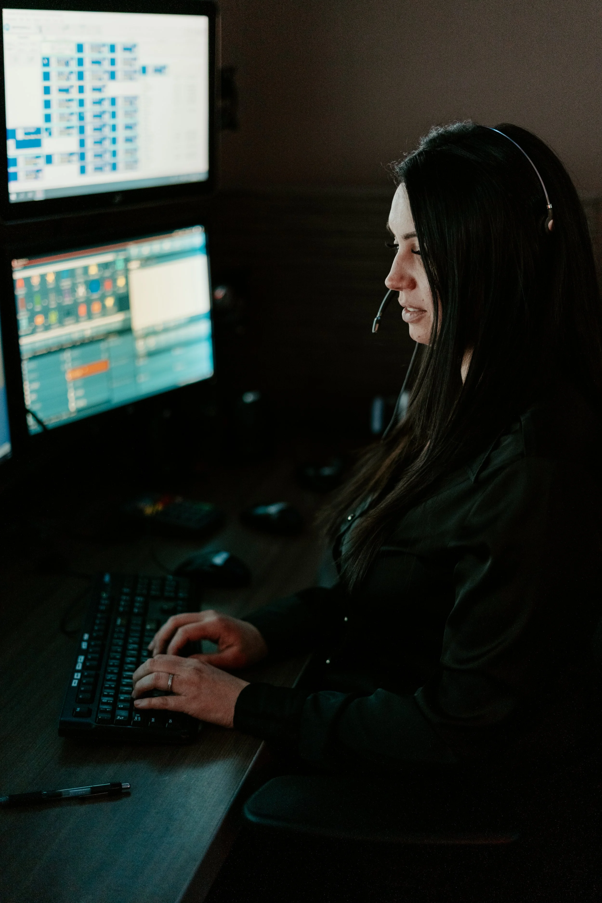 A woman with long dark hair wearing a headset is working at a desk with multiple monitors displaying complex data and charts.