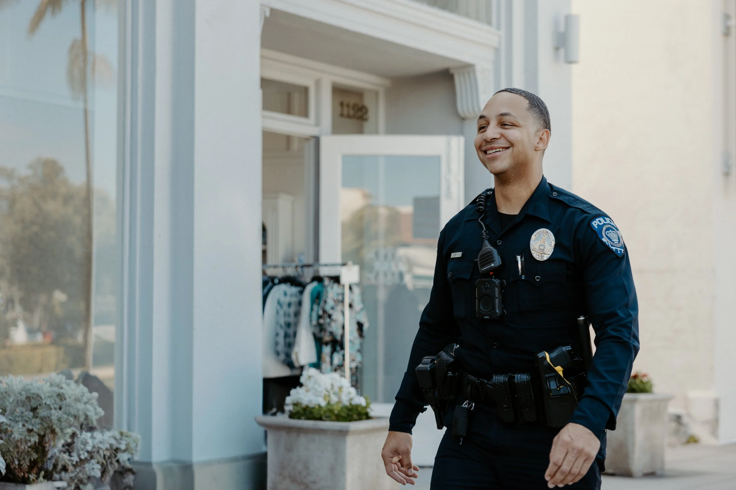 A smiling police officer in uniform walking outside a building, with a flowerpot and clothing rack in the background.