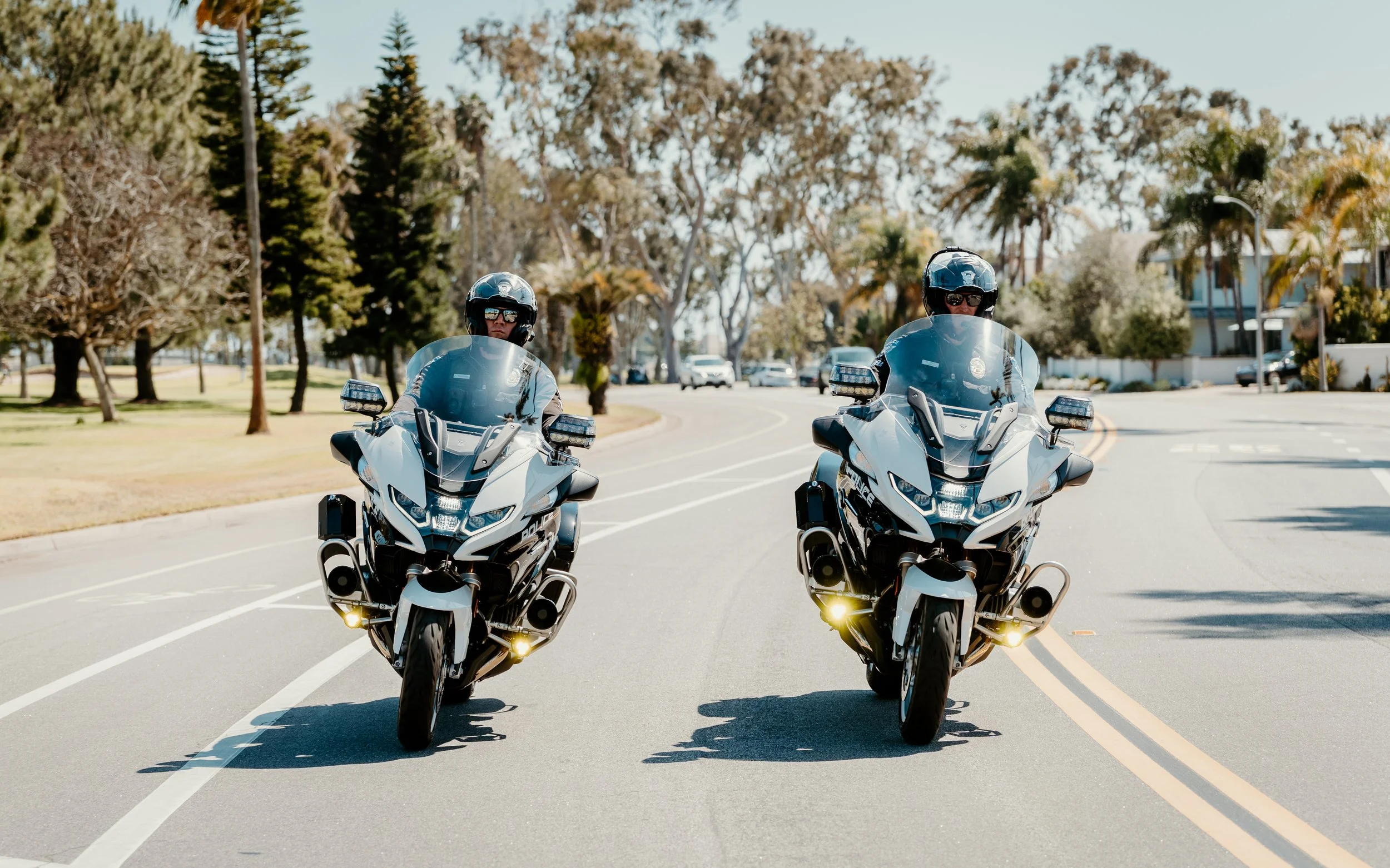 Two police officers riding white police motorcycles on a sunny suburban street with trees and houses in the background.