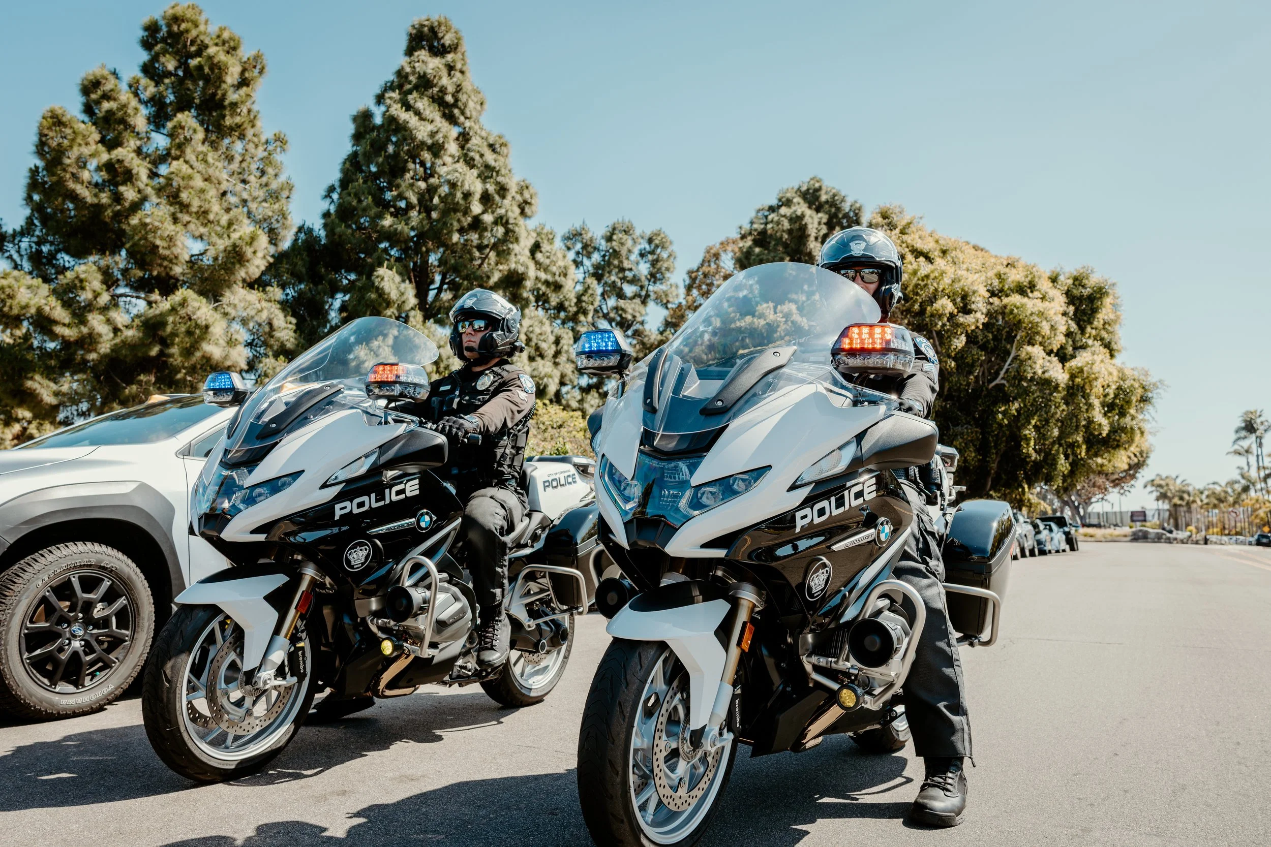 Two police officers on BMW motorcycles parked on the side of a street, with trees and other vehicles in the background.