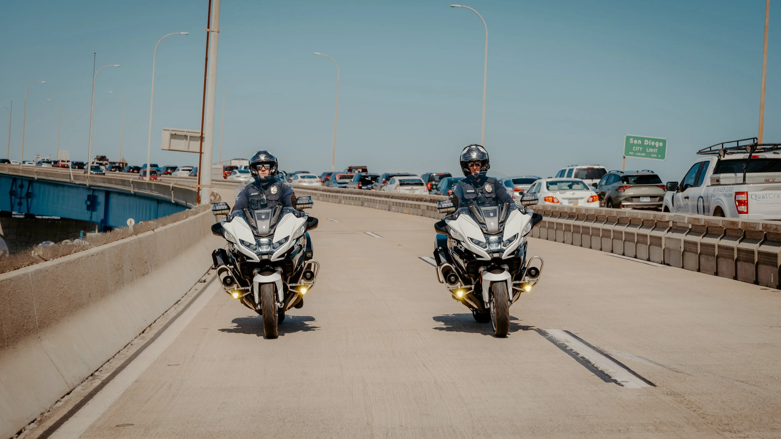 Two police officers riding police motorcycles side by side in the middle of a highway with a busy parking lot on the right and a sign indicating San Diego in the background.