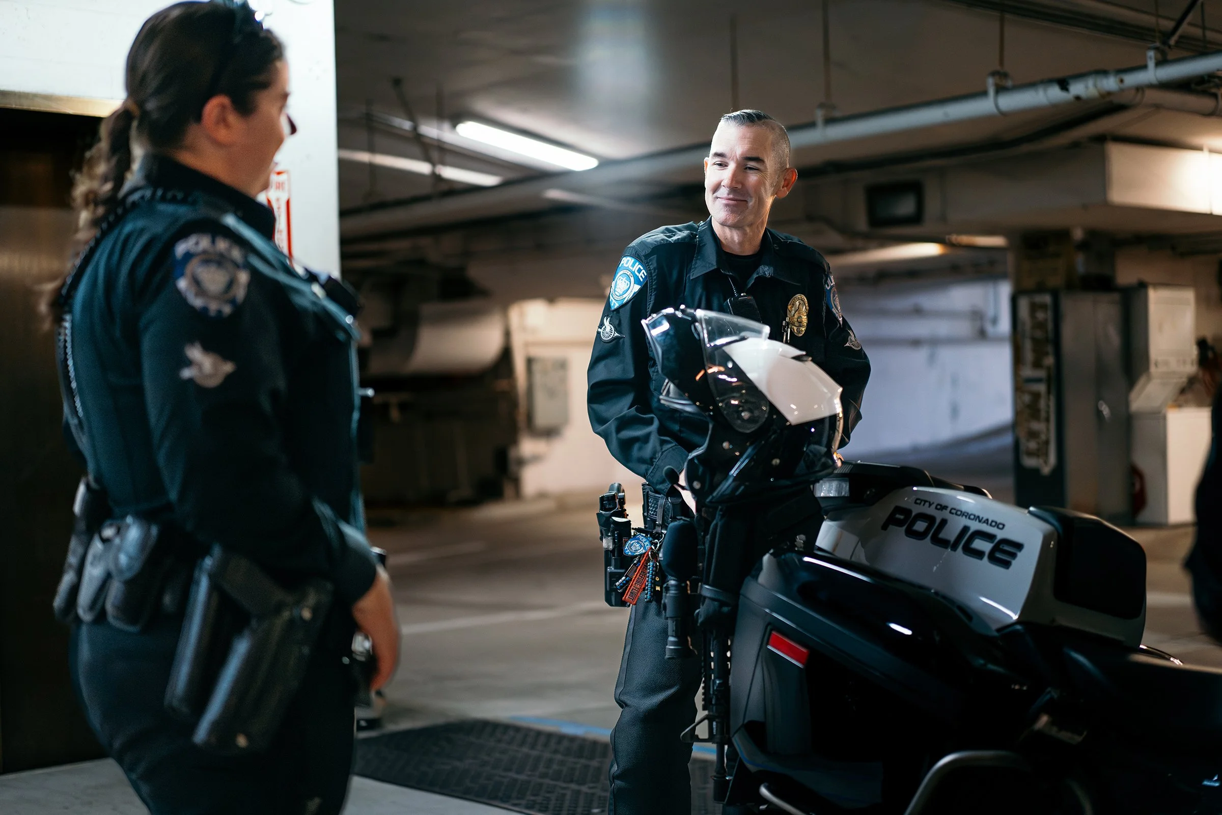 Two police officers, one male and one female, talking in a parking garage. The male officer is leaning on a police motorcycle with a badge and a radio clip, while the female officer is standing nearby.