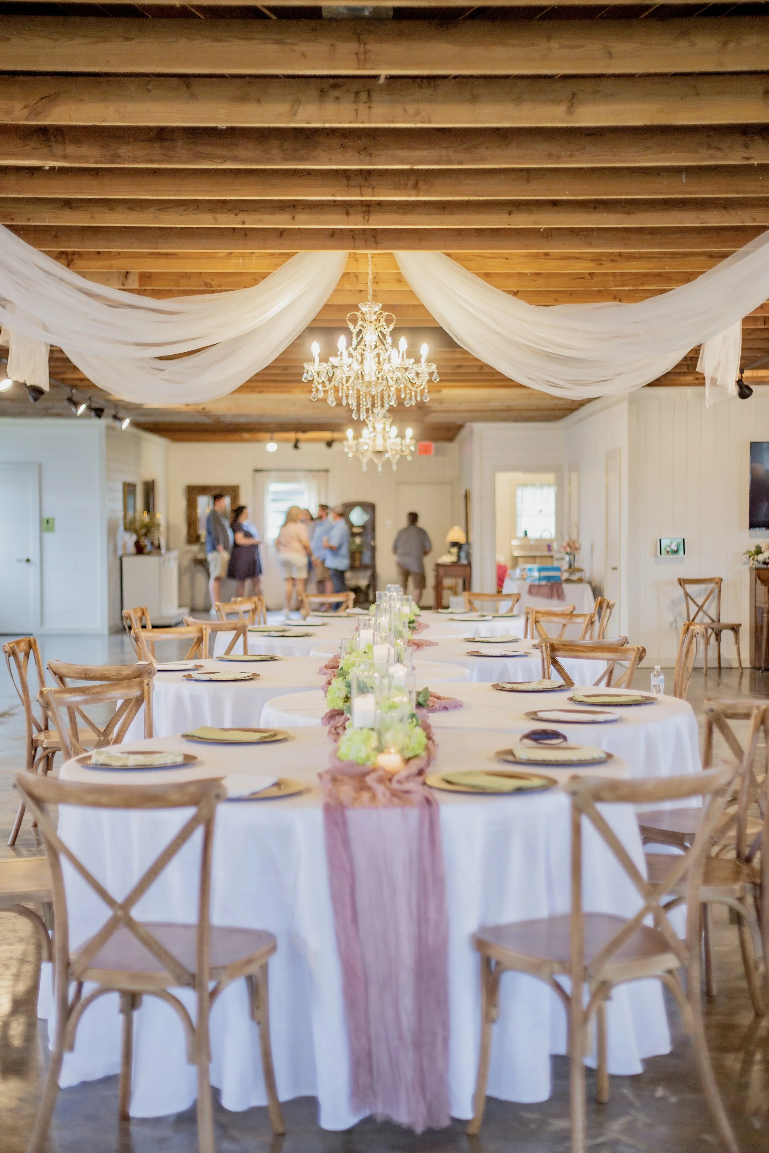 Elegant banquet table setup in a rustic wedding venue with white tablecloths, pink and green floral centerpiece, candles, wooden chairs, draped fabric, and chandelier ceiling decoration, with people in the background.