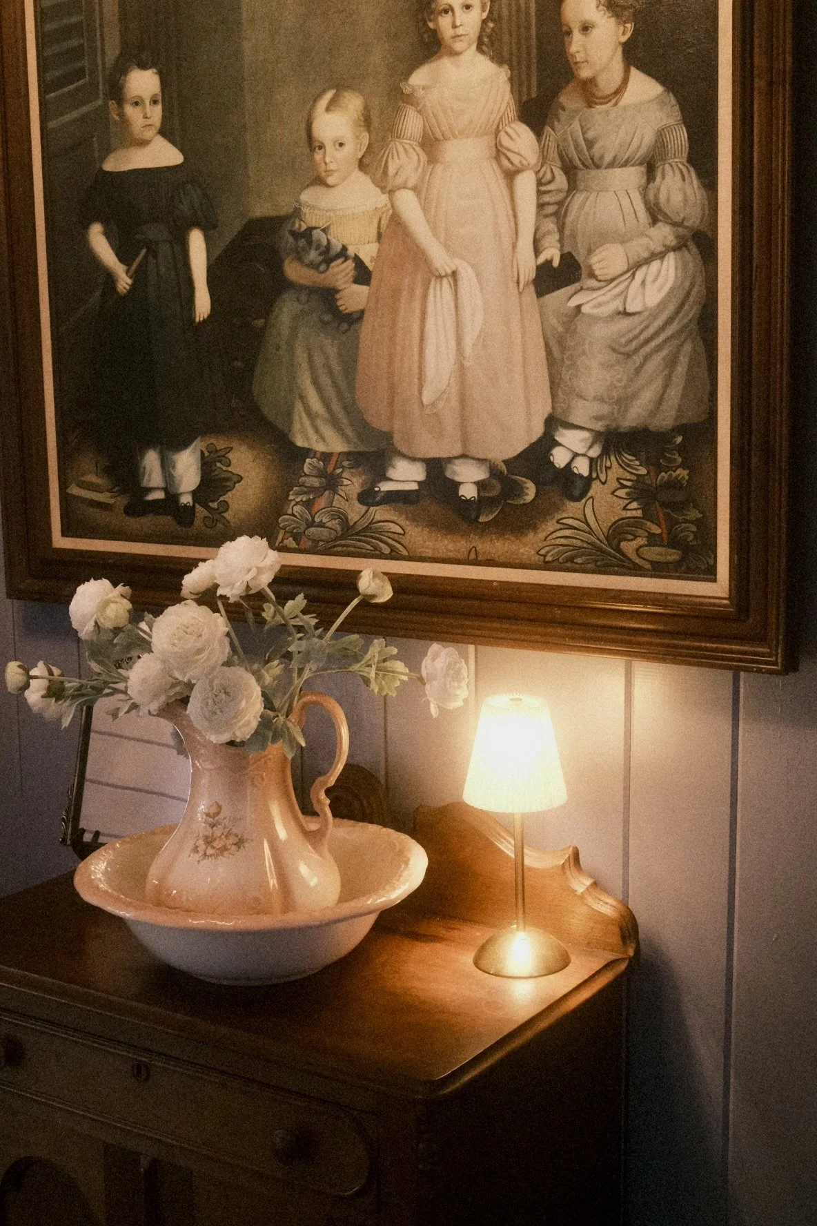 Painting of five children dressed in historical clothing hanging on the wall, with a table below holding a vase of white flowers and a small lamp.