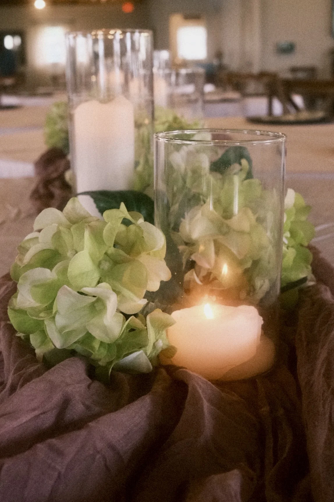Decorative centerpiece featuring a lit candle in a glass holder, surrounded by hydrangea flowers and greenery, arranged on a table with a brown tablecloth.
