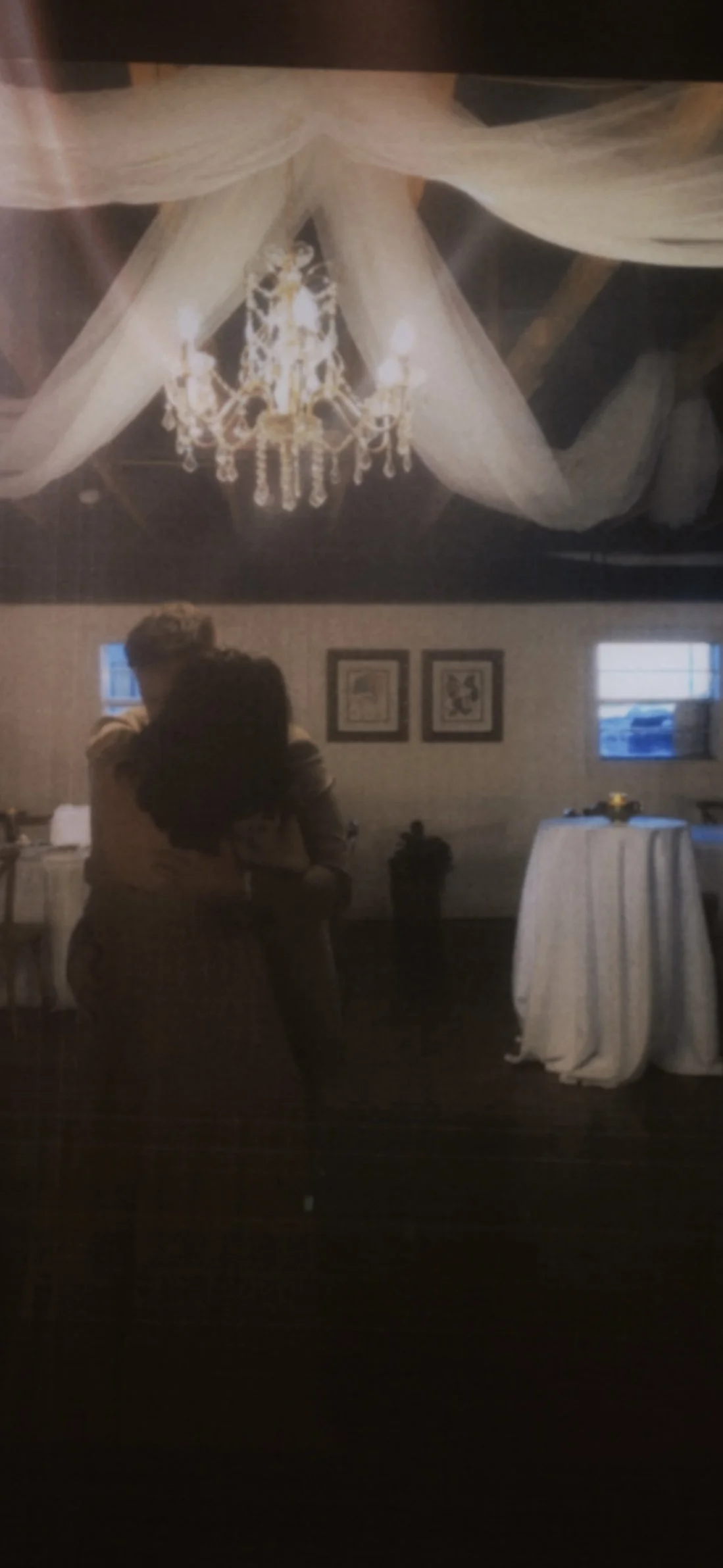 A couple embraces in a dimly lit room with draped ceiling fabric, a chandelier, framed pictures on the wall, windows, and tables covered with white tablecloths.