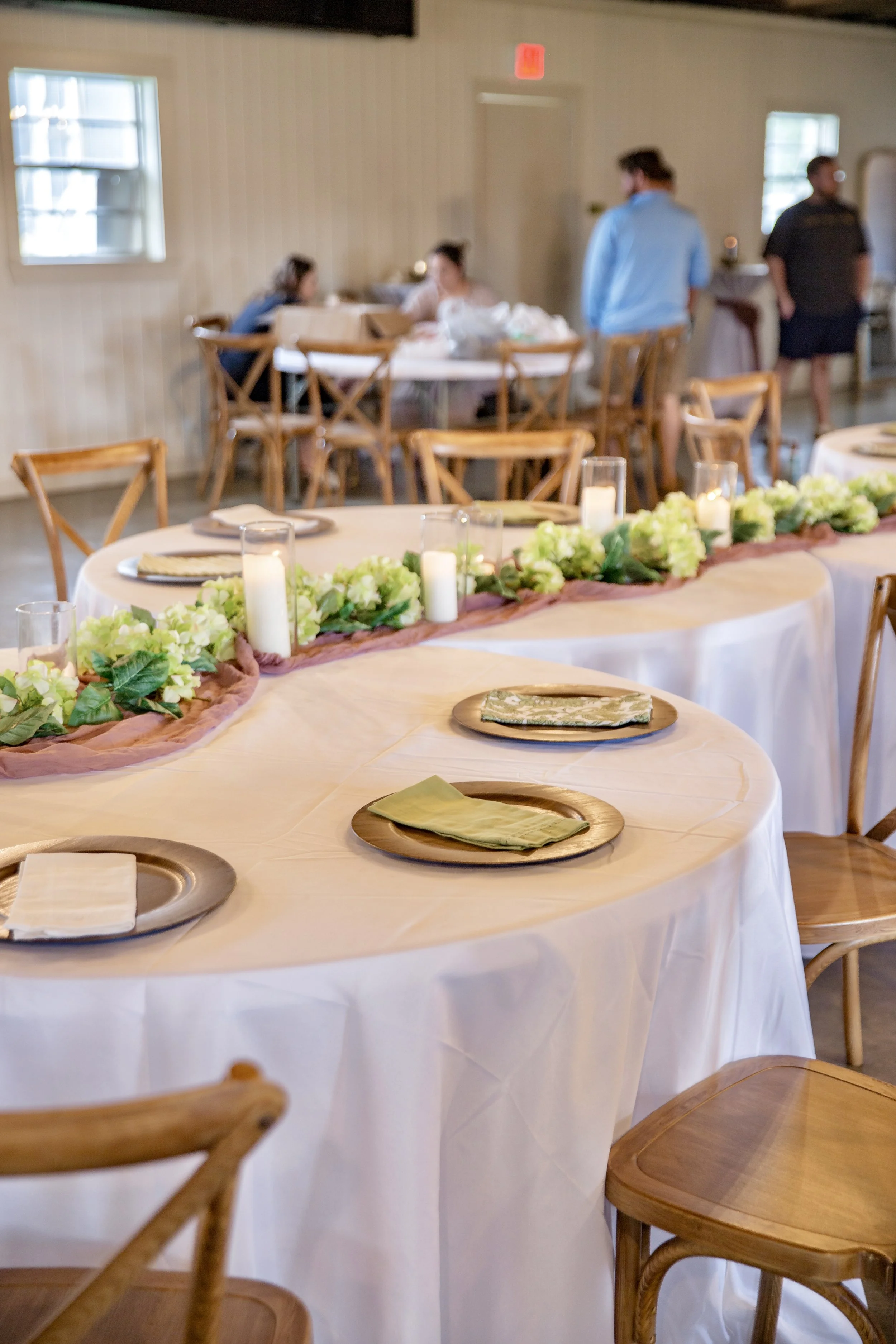 Round tables with white tablecloths and a decorative runner with green and white floral arrangements and candles. Wooden chairs around the tables, in a well-lit room with windows on the walls. People in the background preparing for an event or gather
