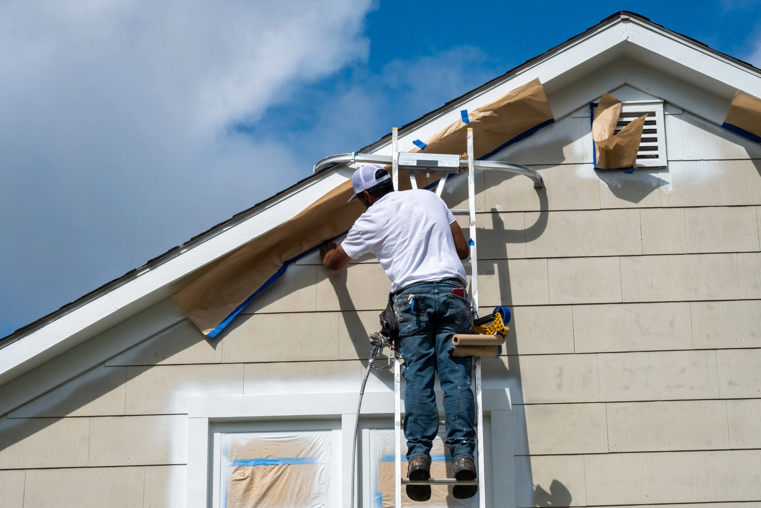 A construction worker in a white t-shirt and jeans is standing on a ladder installing or repairing the soffit and fascia at the edge of a house roof. The house has cream-colored siding, and parts are covered with brown paper for protection, with some blue tape securing it. The worker is facing away from the camera, working on the house exterior against a partly cloudy sky.