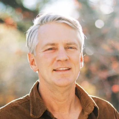A middle-aged man with light gray hair in an outdoor setting with blurred trees and autumn leaves.