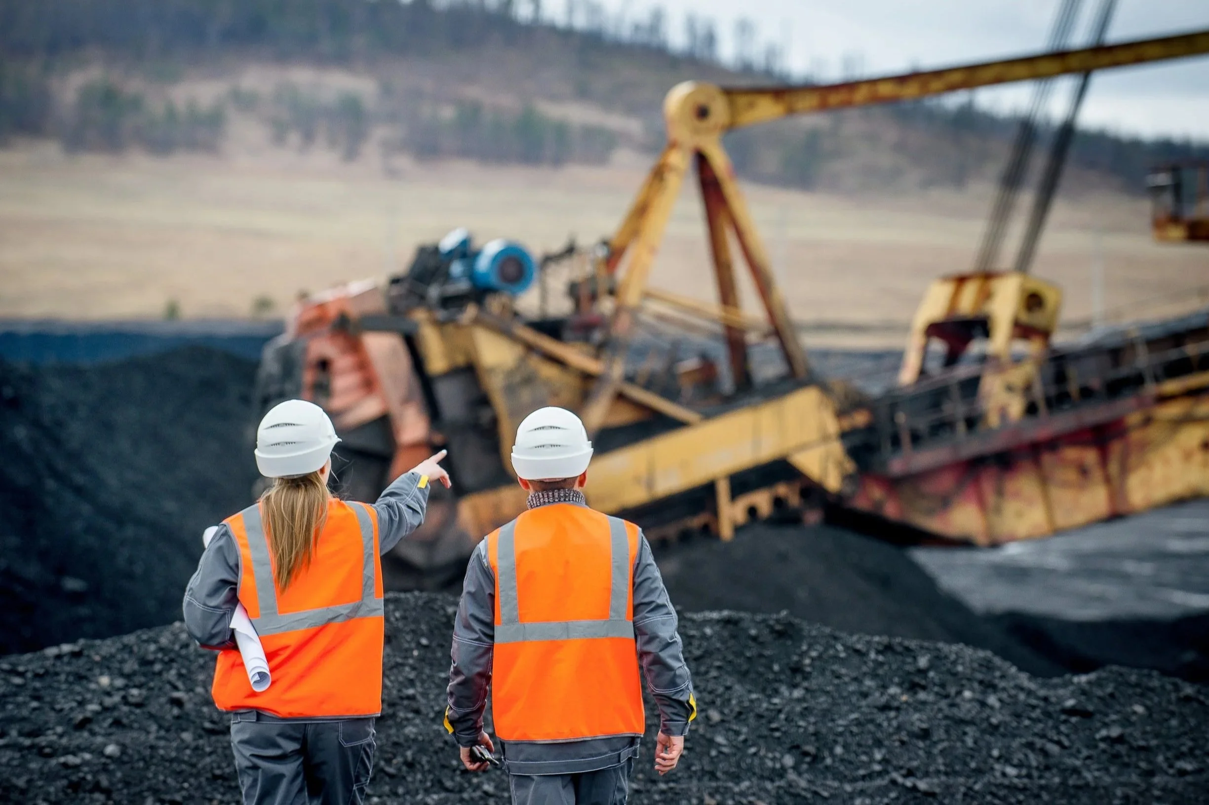 Mining workers wearing orange PPE and white helmet with an excavator in the background
