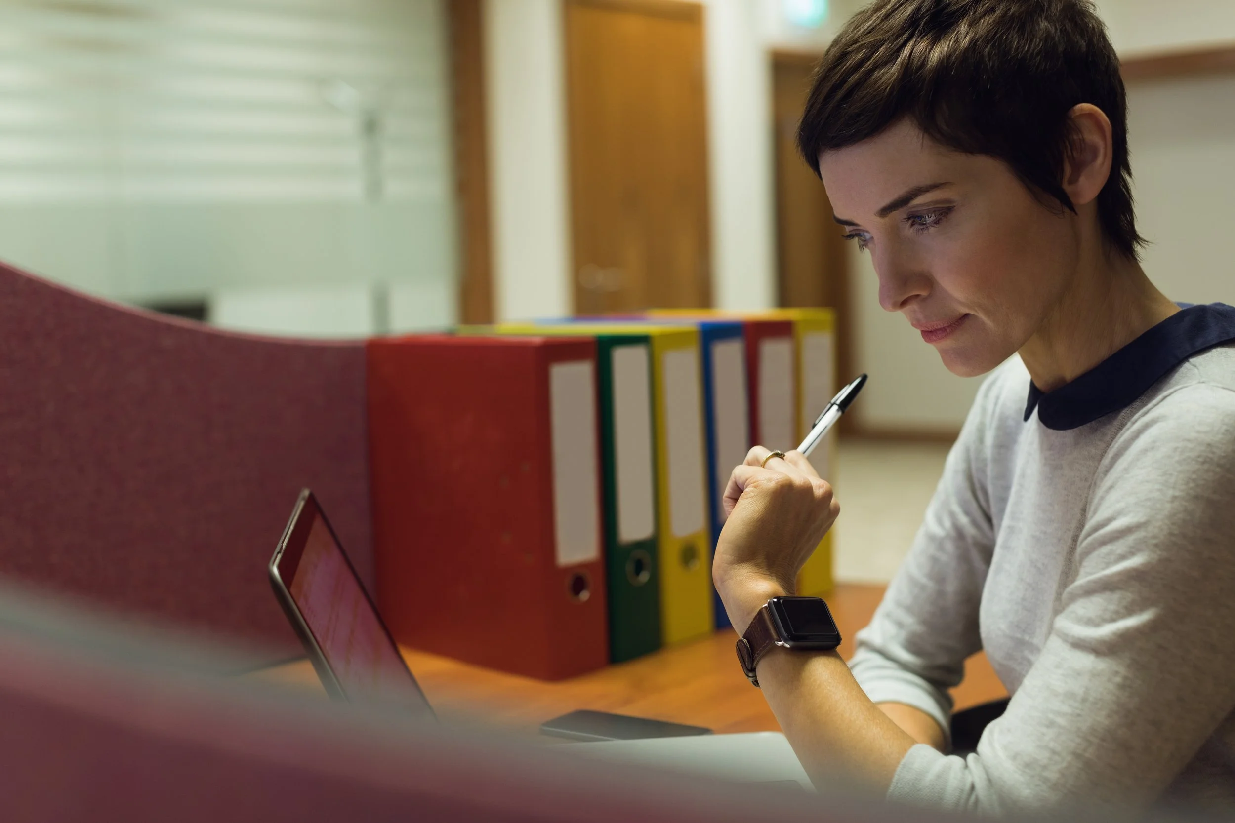 Research Studies represented by a woman wearing a white shirt with a black collar, sitting at a desk with colorful binders, looking at her smartphone and holding a pen.