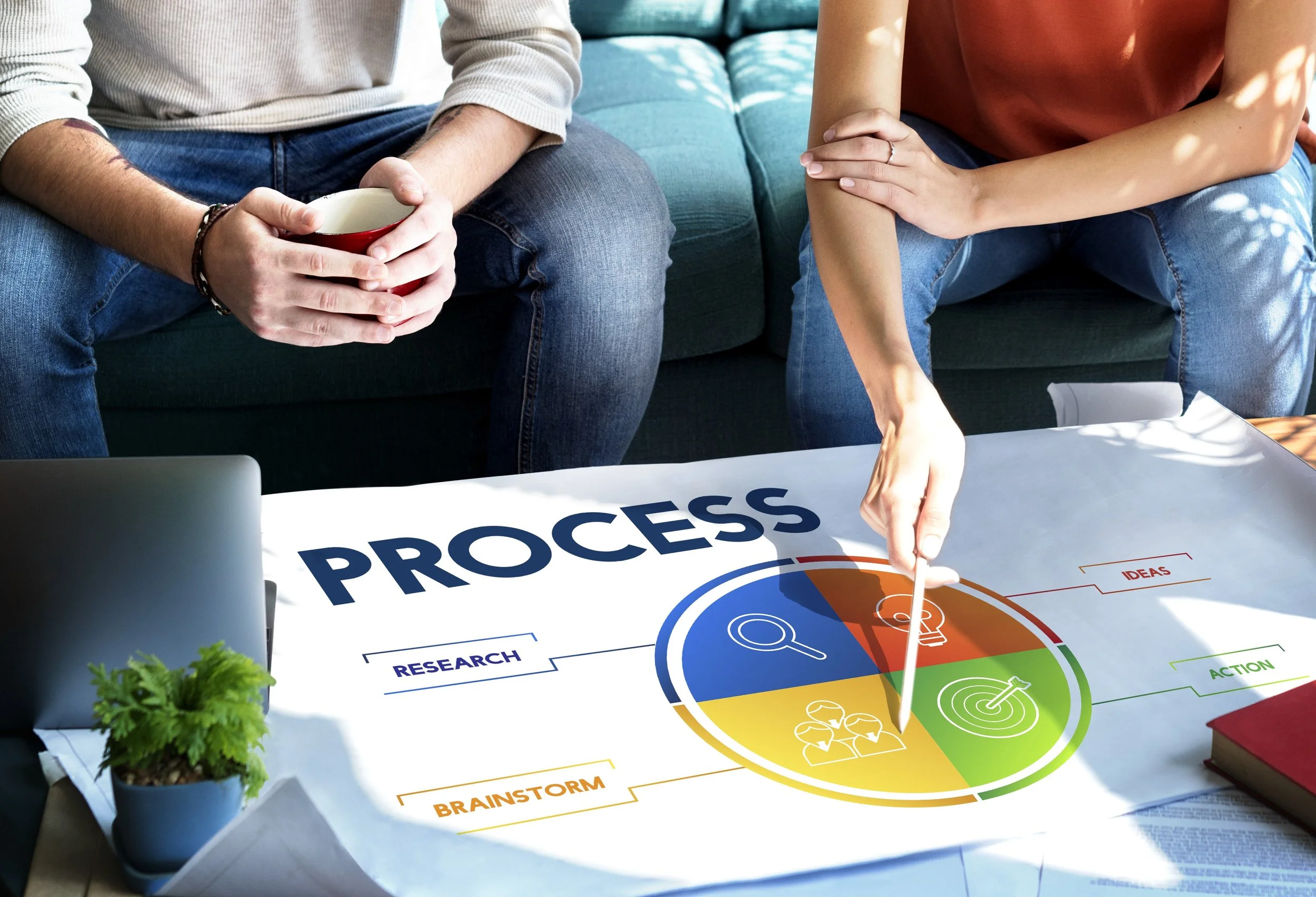 Two people sitting at a table with a process diagram on paper, one pointing at it with a pen and the other holding a coffee mug, in a casual office setting.