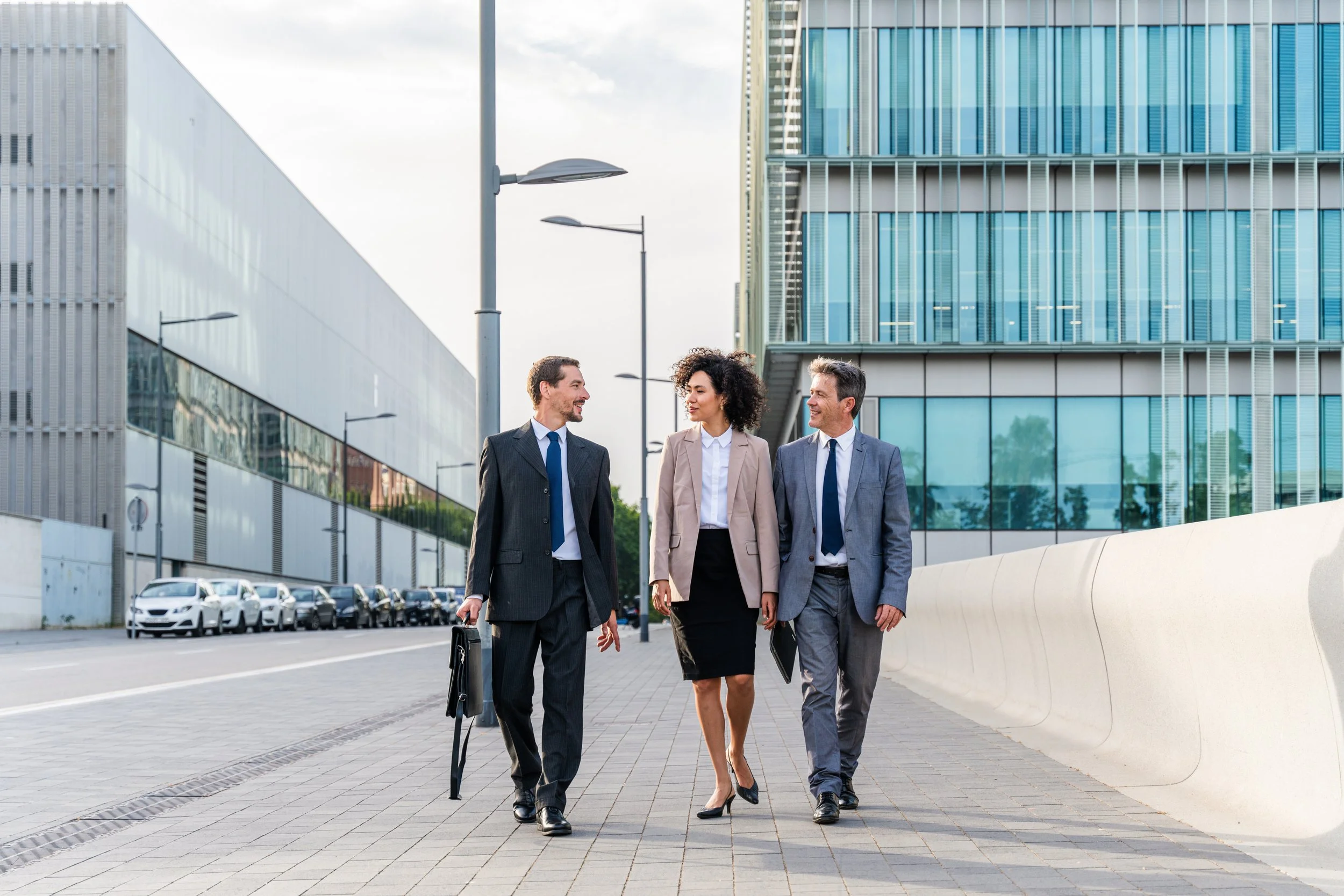Three business professionals walking and talking outside in an urban area with modern glass buildings.