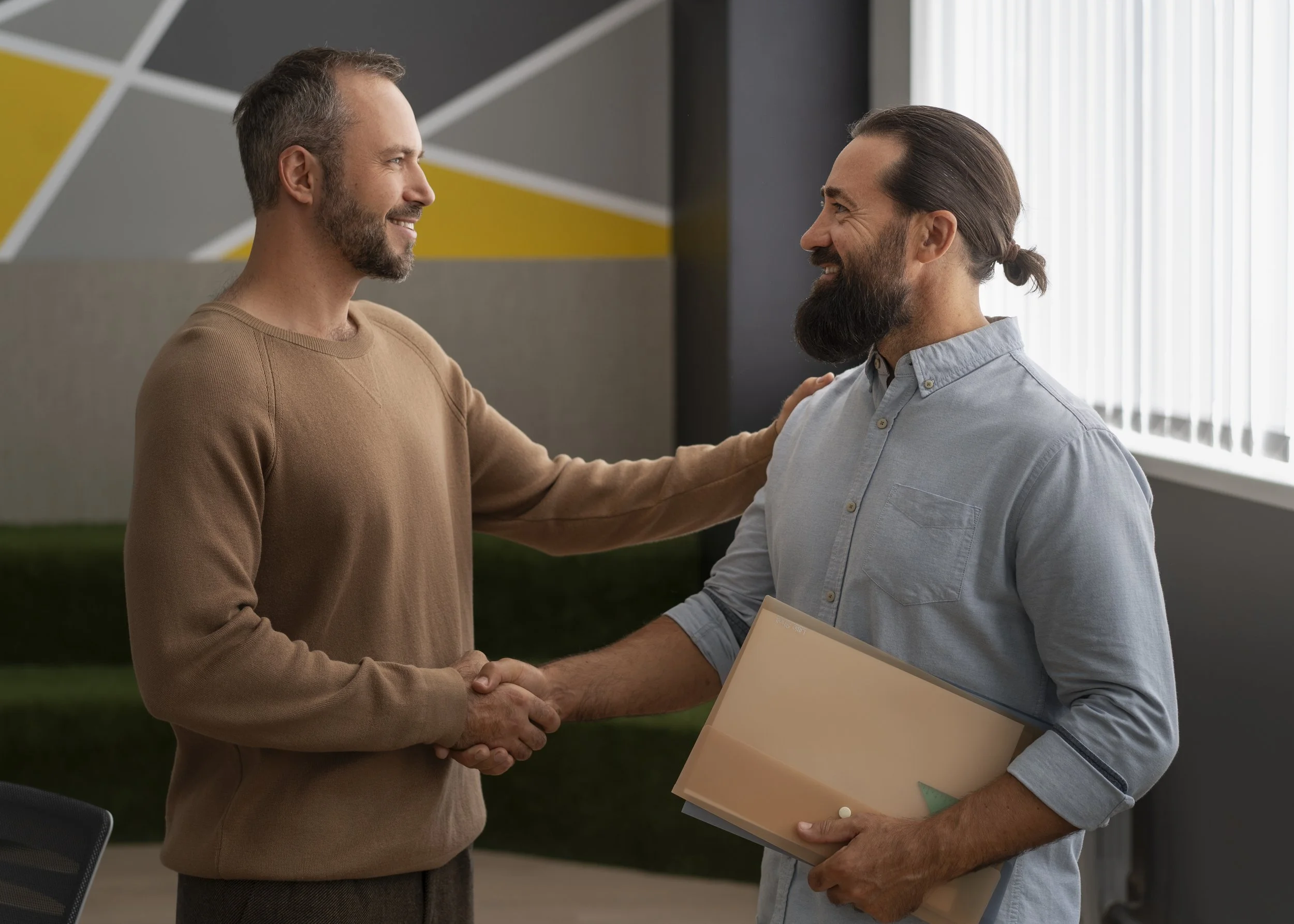 Two men shaking hands in an office, both smiling, one holding folders, the other affectionately touching the other's shoulder.