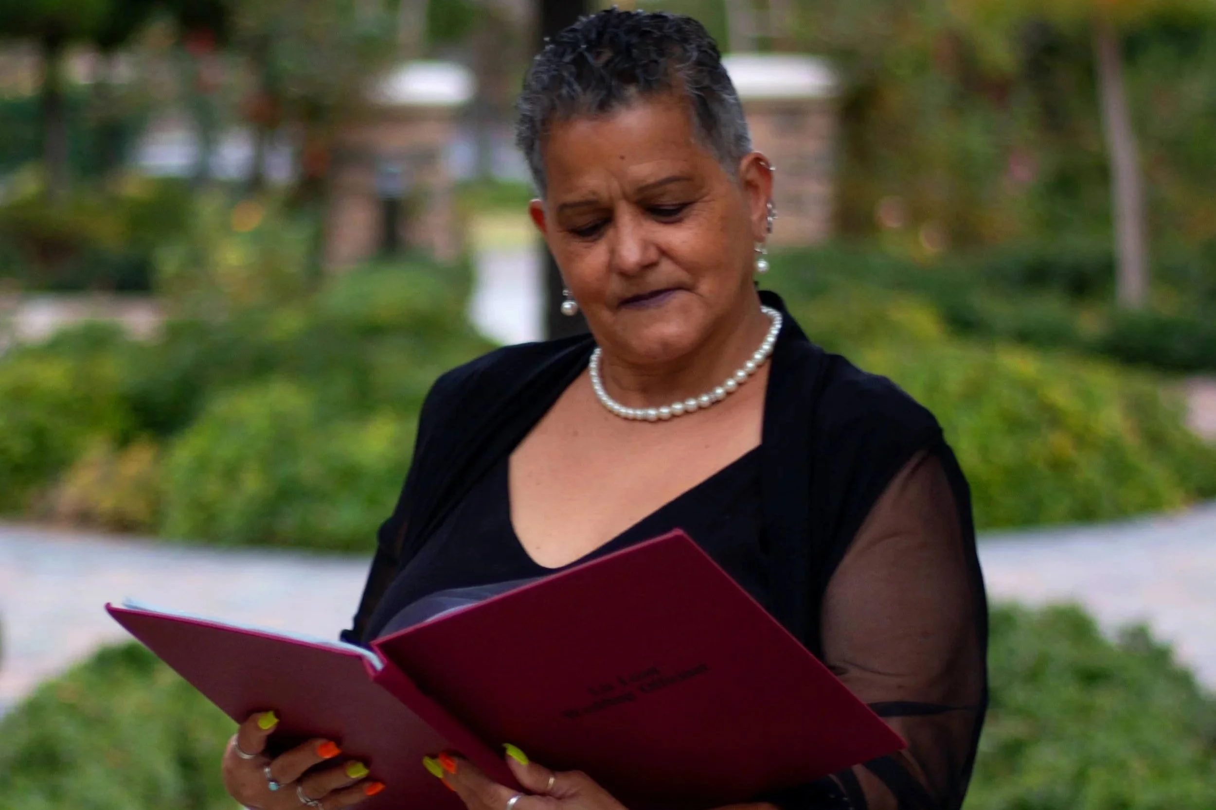 Woman with short gray hair and pearl necklace reading a pink booklet outdoors with trees in the background.