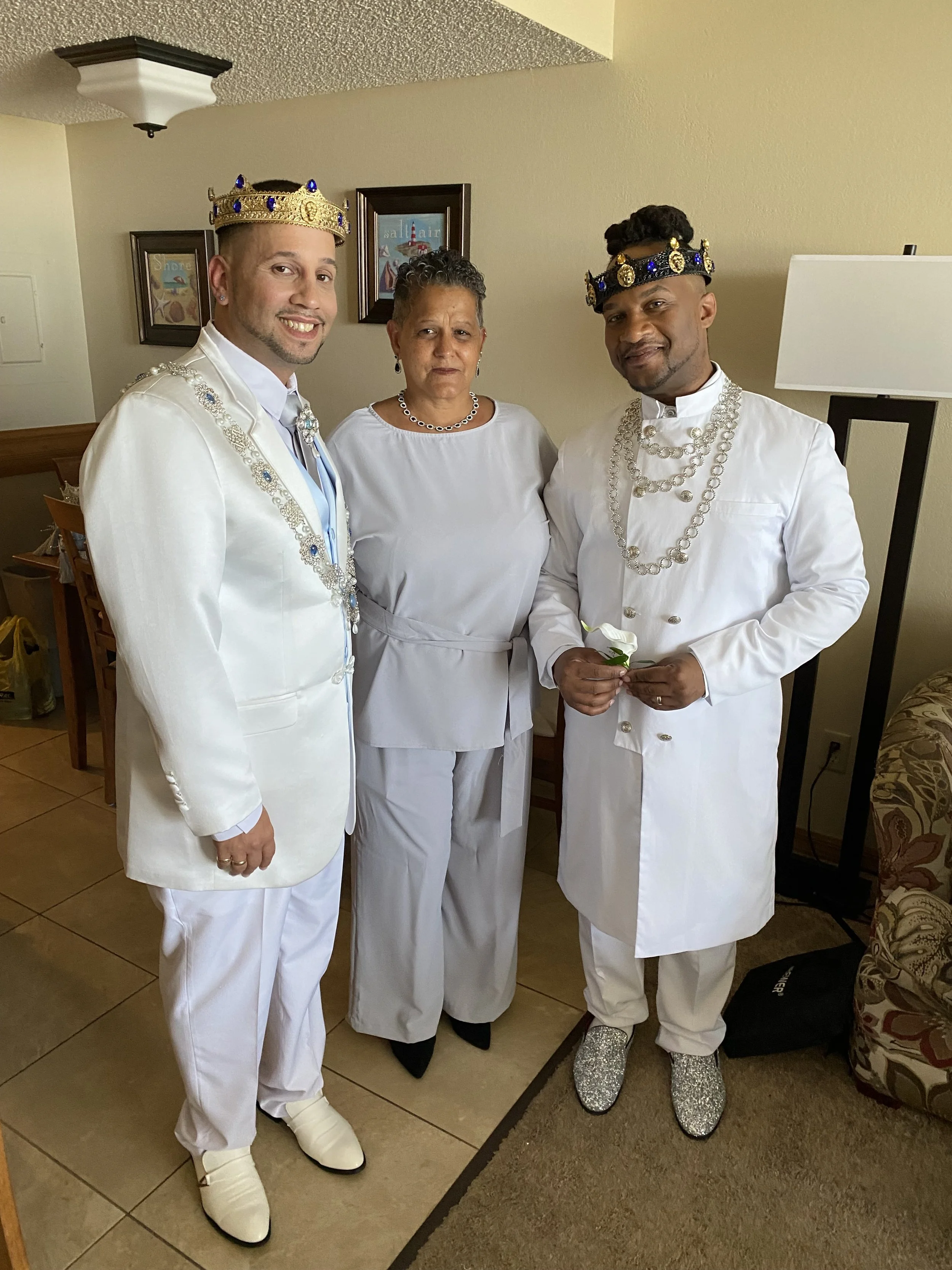 Two men wearing traditional Indian wedding attire, holding candles and candles in front of a lit candle and cream-colored curtains.