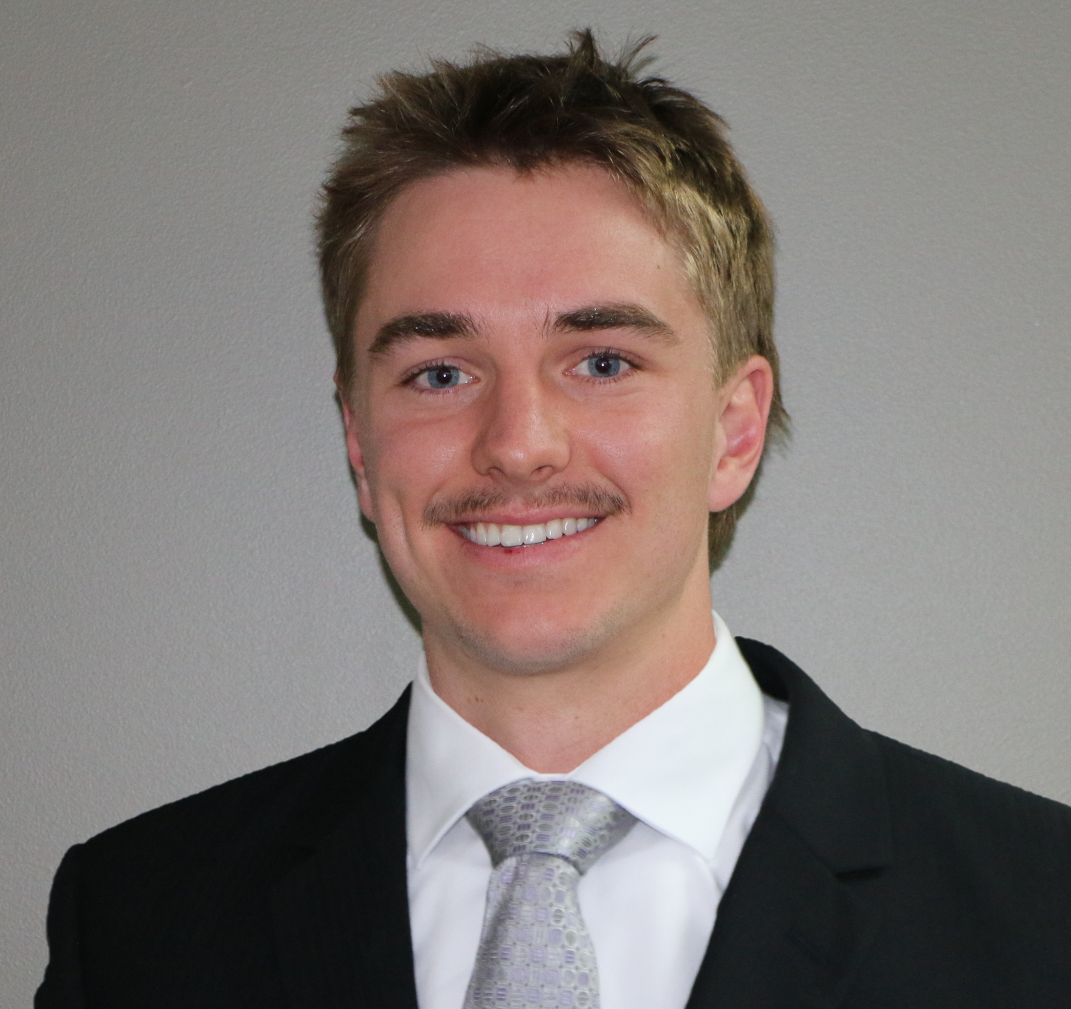 Portrait of a young man with light brown hair, blue eyes, and a mustache, wearing a black suit, white shirt, and light gray patterned tie, smiling in front of a plain gray wall.