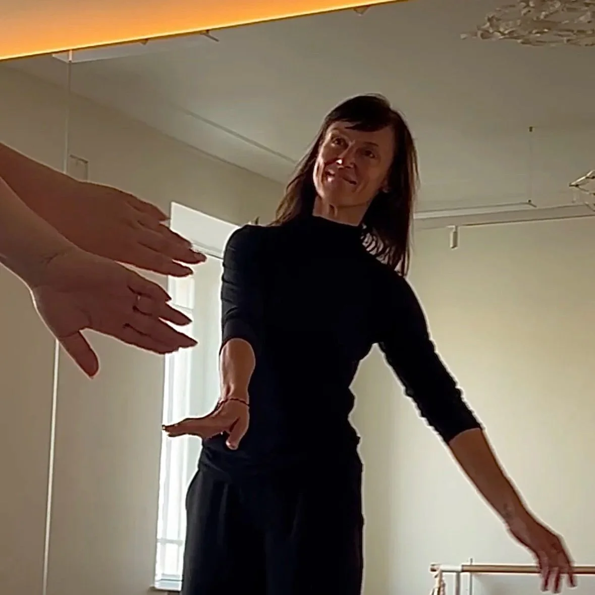 A smiling woman in a black long-sleeve top is reaching out with her right hand toward a group of hands extended towards her. She is indoors, with a window and ceiling lights in the background.