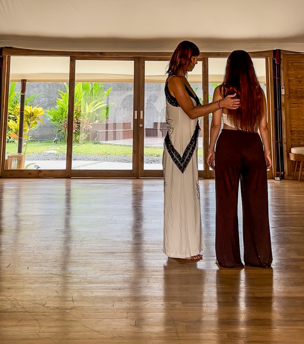 A woman with brown hair in a black top and pants standing and smiling while reaching out her hand in a dance studio with a mirror and ballet bar.