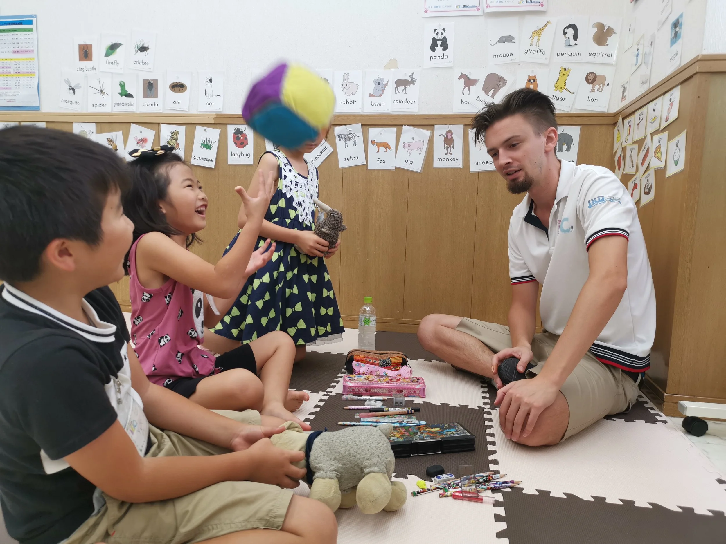 子供たちが室内で遊びながら笑っている。先生が座っている。カラフルなボール、ぬいぐるみ、絵具、文房具が床に置かれている。壁には動物の絵と名前のカードが貼られている。