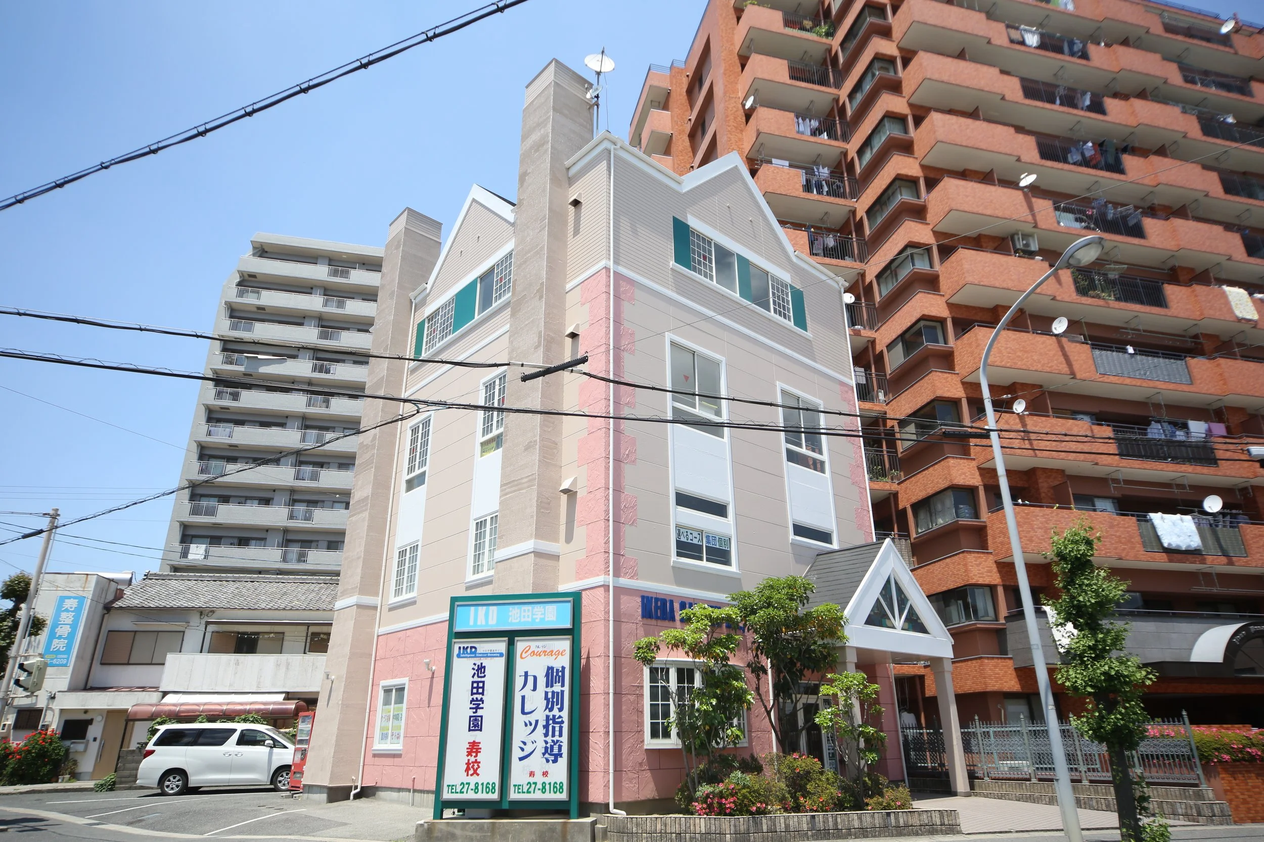 ひとつの白とピンクの建物と、その前には木があり、看板があります。背景には高いマンションが建っています。