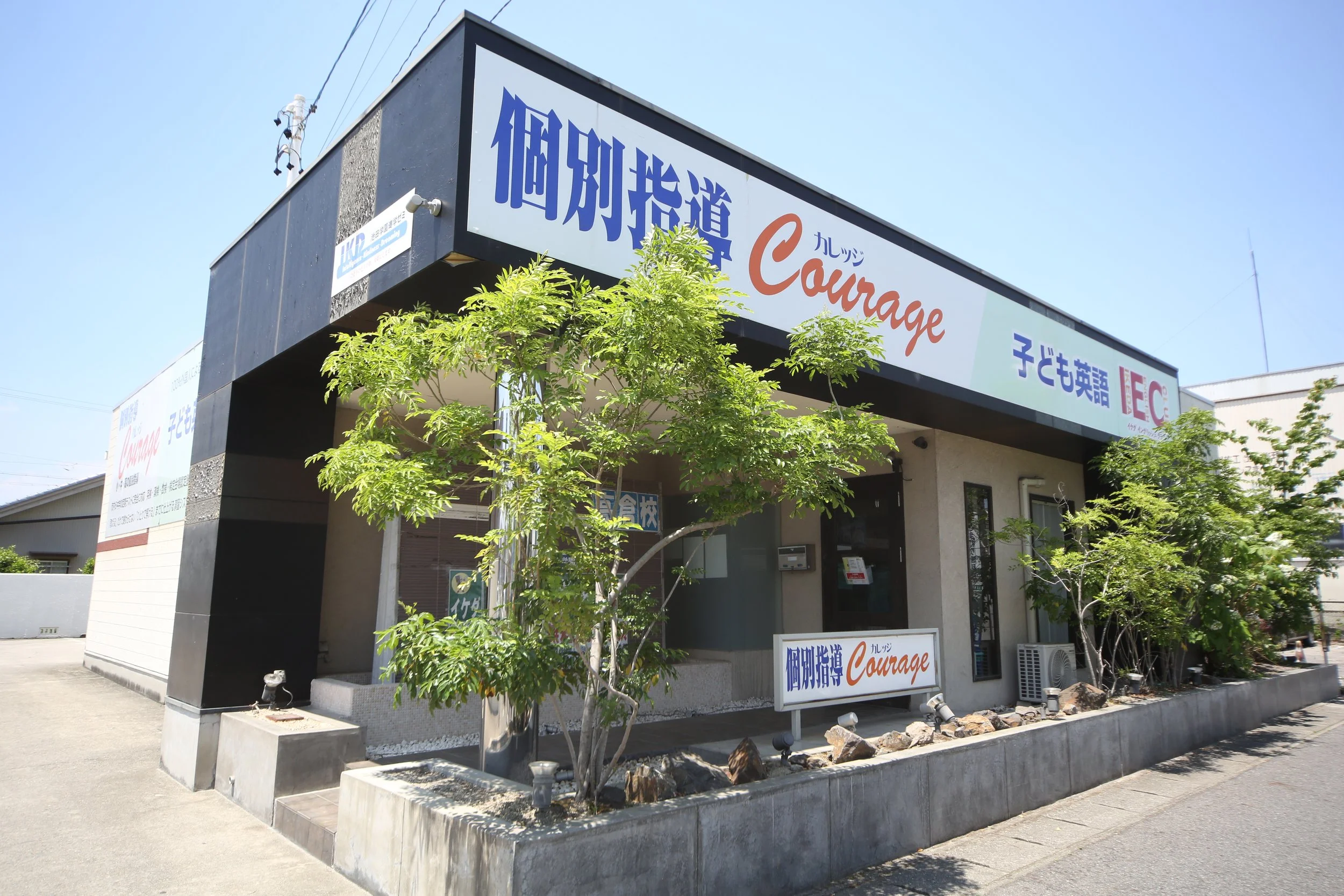 黒い建物の外観、複数の木と看板があり、看板には日本語と英語で「個別指導Courage」と書かれている。