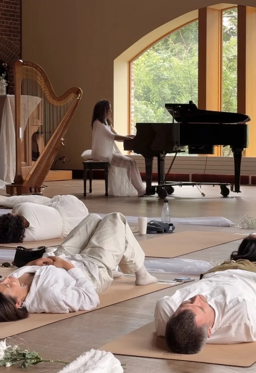 Woman playing the grand piano during a yoga or meditation session, with people lying on yoga mats in the foreground inside a room with large windows showing greenery outside.