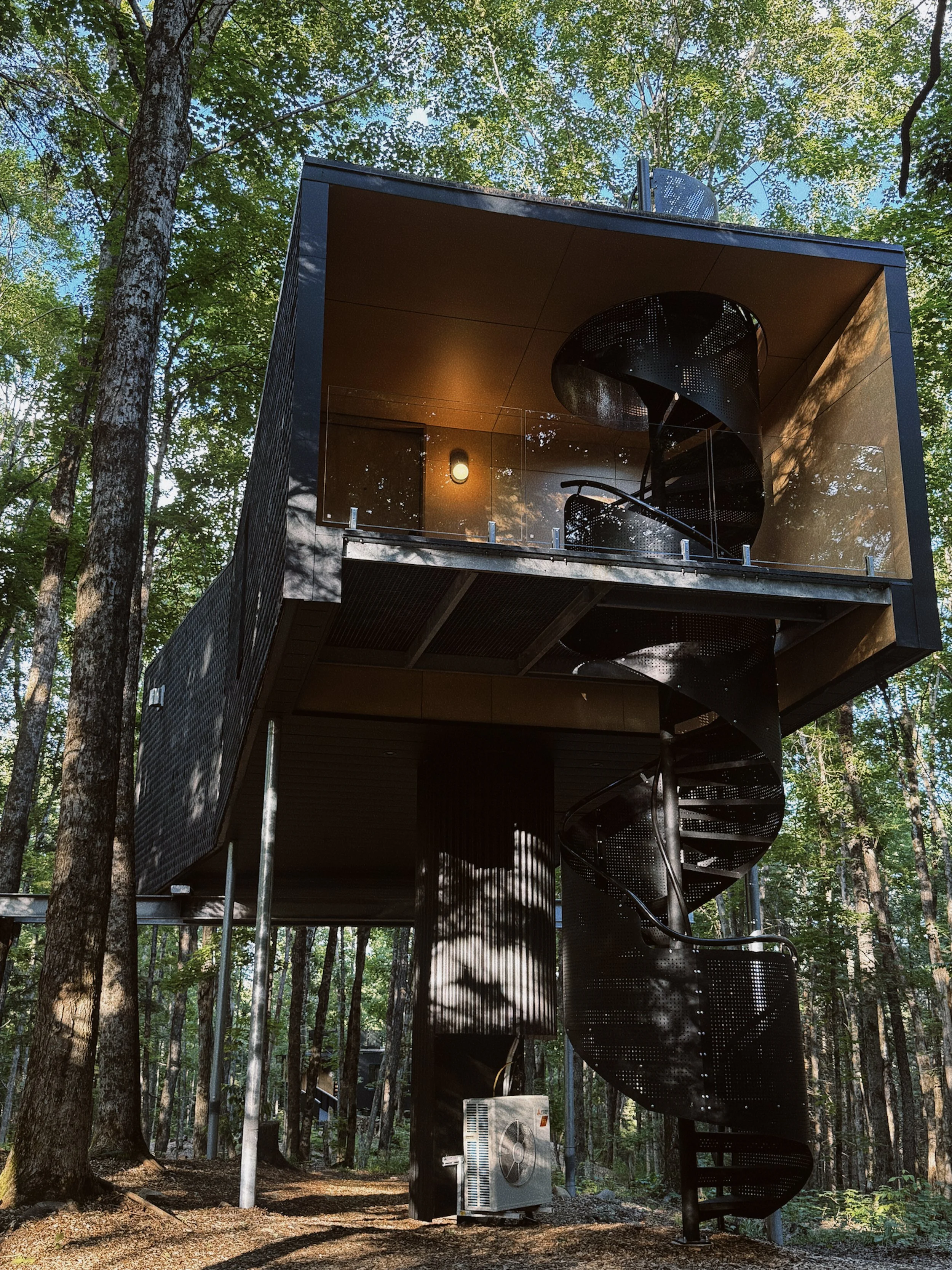A modern, black, multi-story building with outdoor spiral staircase in a forest setting. The building has glass railings on the balcony and is surrounded by tall trees with green leaves and a partly cloudy sky.