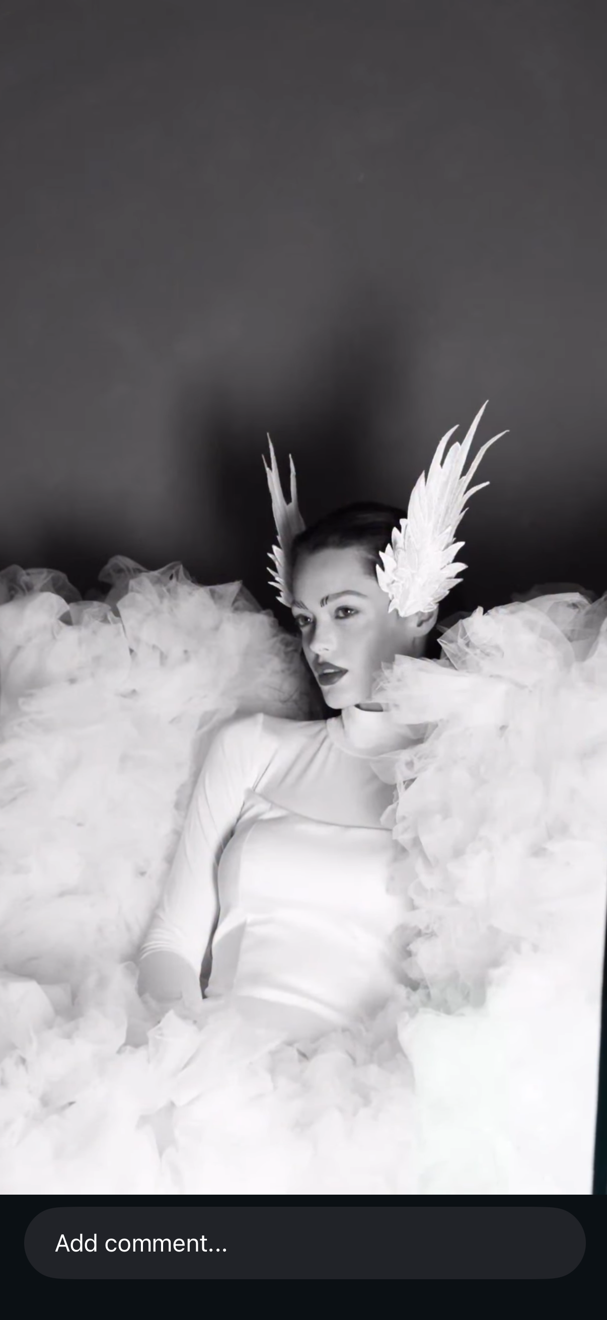 A woman with a shaved head wearing large feathered earrings and surrounded by fluffy tulle, posing in a dramatic fashion against a dark background.