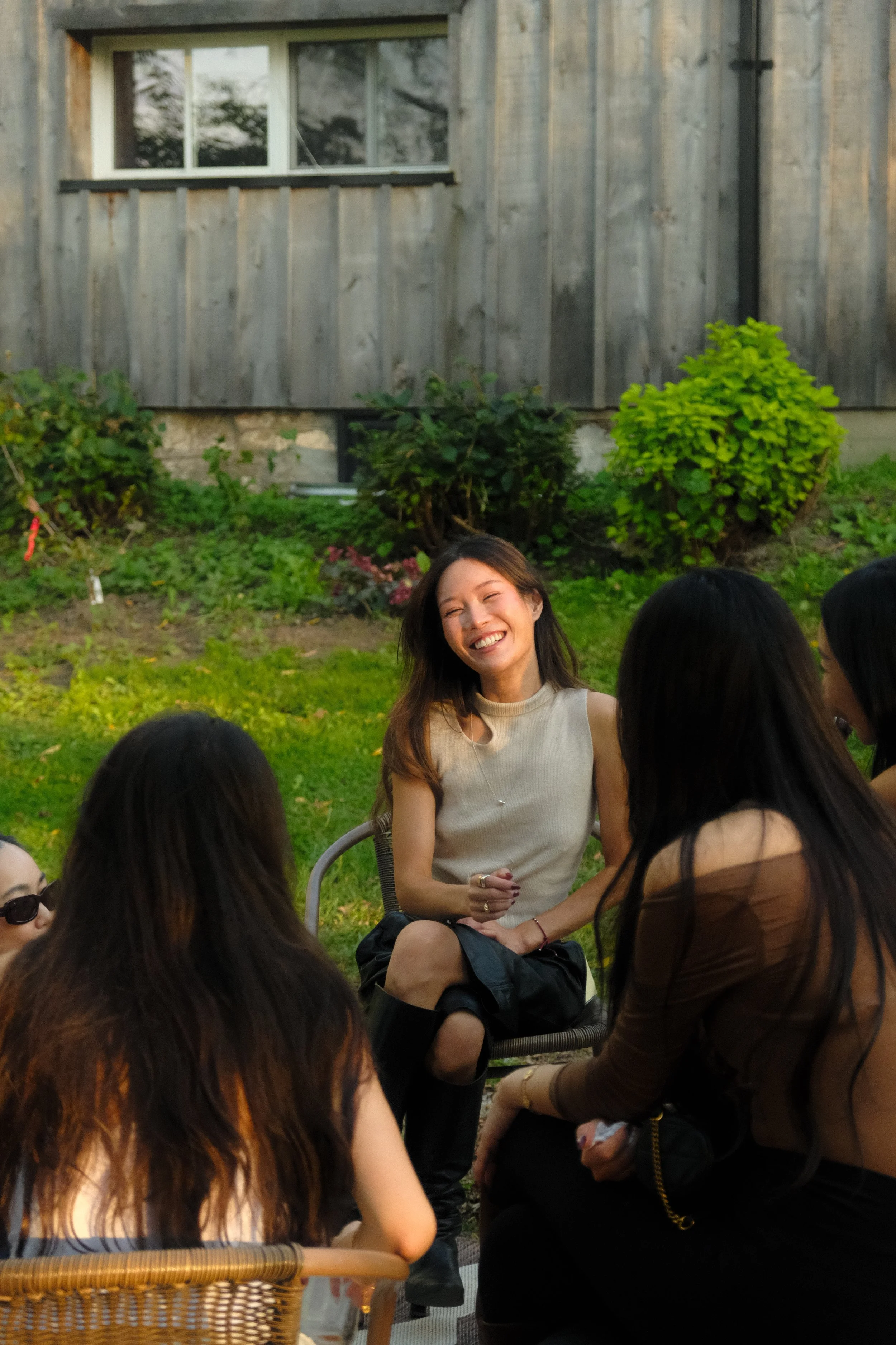 A group of people gathered outdoors in a garden with green plants and bushes, engaging in conversation and smiling. A woman with long brown hair, wearing a sleeveless top, is the focus of the image.