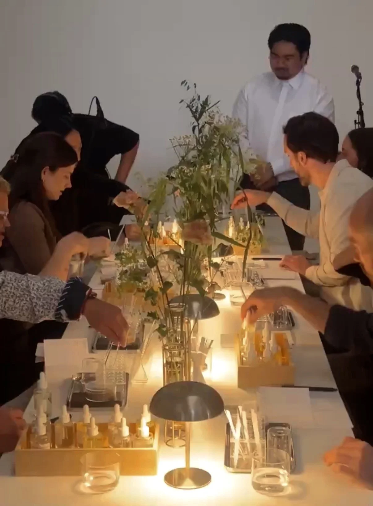 A group of people gathered around a table lighting candles, with a tall plant centerpiece in the middle and a plain white wall background.