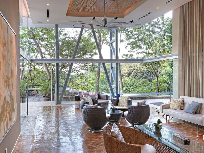 Modern living room with large floor-to-ceiling windows, trees visible outside, and contemporary furniture including sofas, armchairs, a coffee table, and decorative pillows.