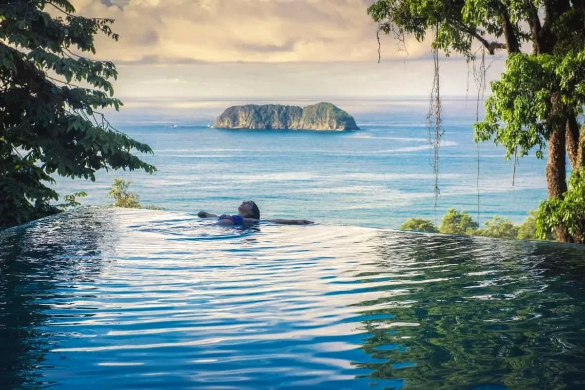 A person floating in an infinity pool overlooking the ocean with a distant island on the horizon, surrounded by trees and a cloudy sky.
