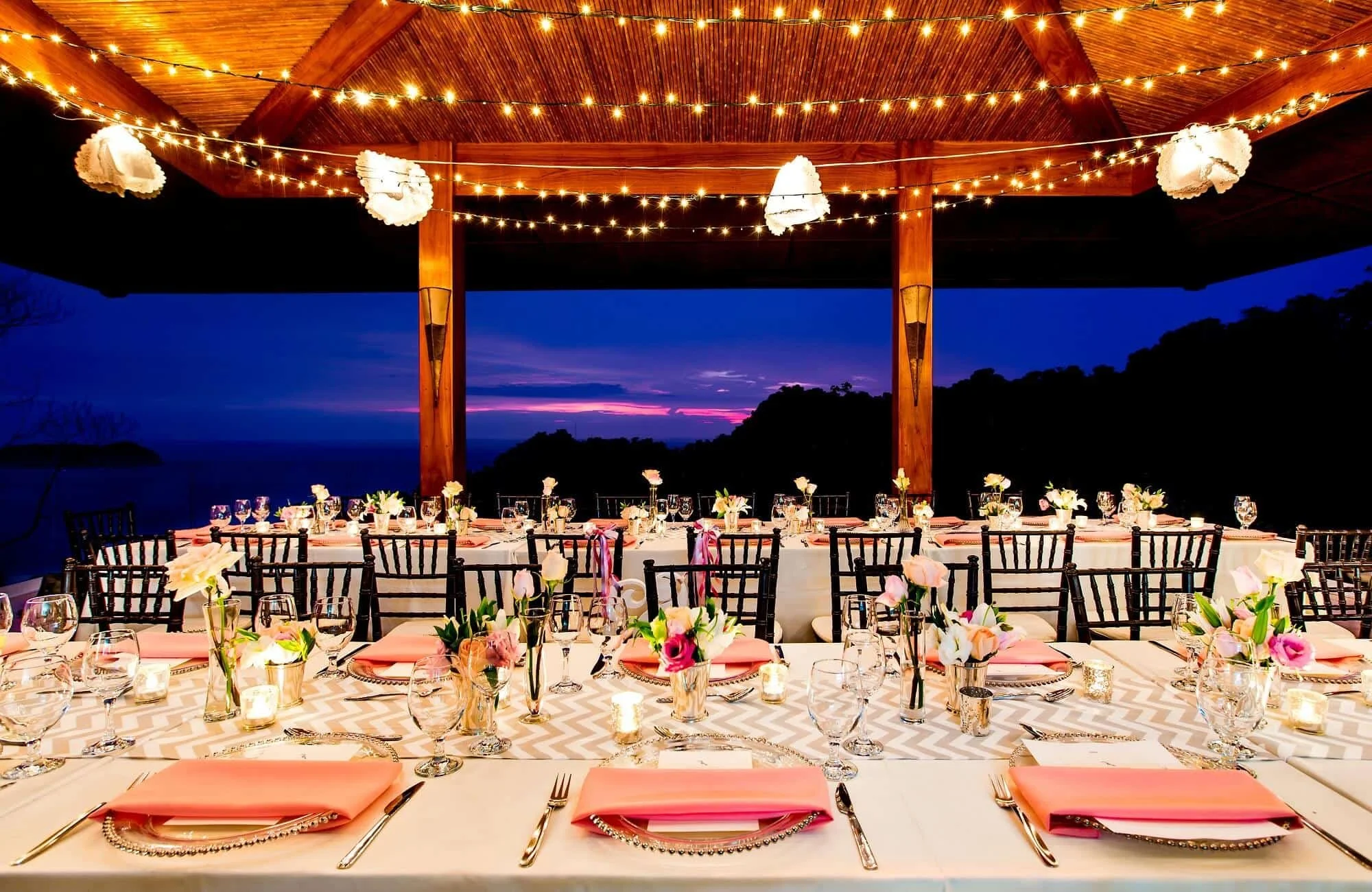 Elegant outdoor dinner setup under a wooden pavilion at dusk. Long tables are decorated with pink napkins, floral centerpieces, candles, and glassware. String lights hang from the ceiling, creating a warm ambiance, with a scenic view of the ocean and a purple sunset in the background.
